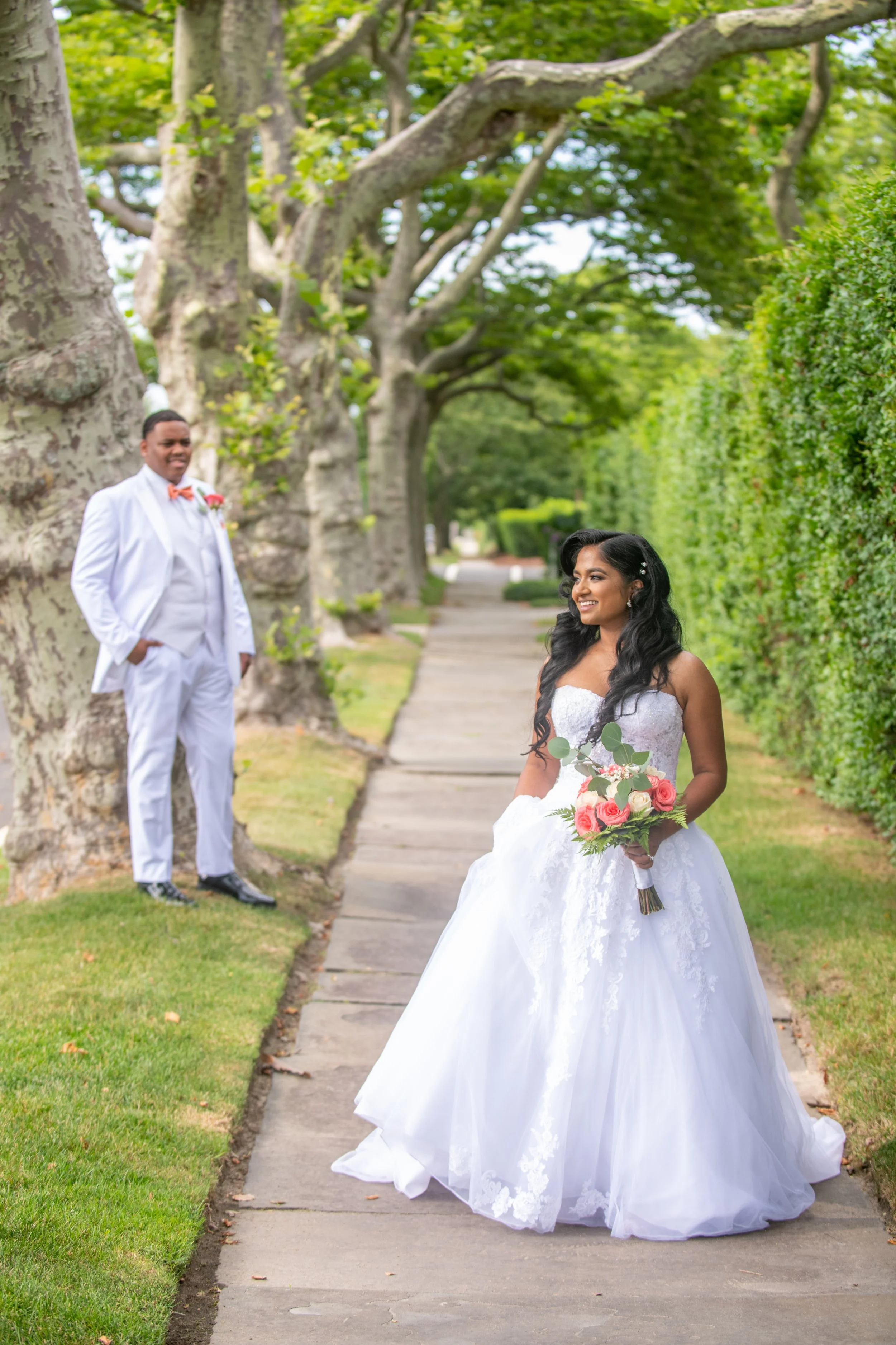 Bride in a white wedding dress holding a bouquet of pink roses and greenery, smiling at groom standing in a white suit with orange bow tie, on a tree-lined sidewalk.