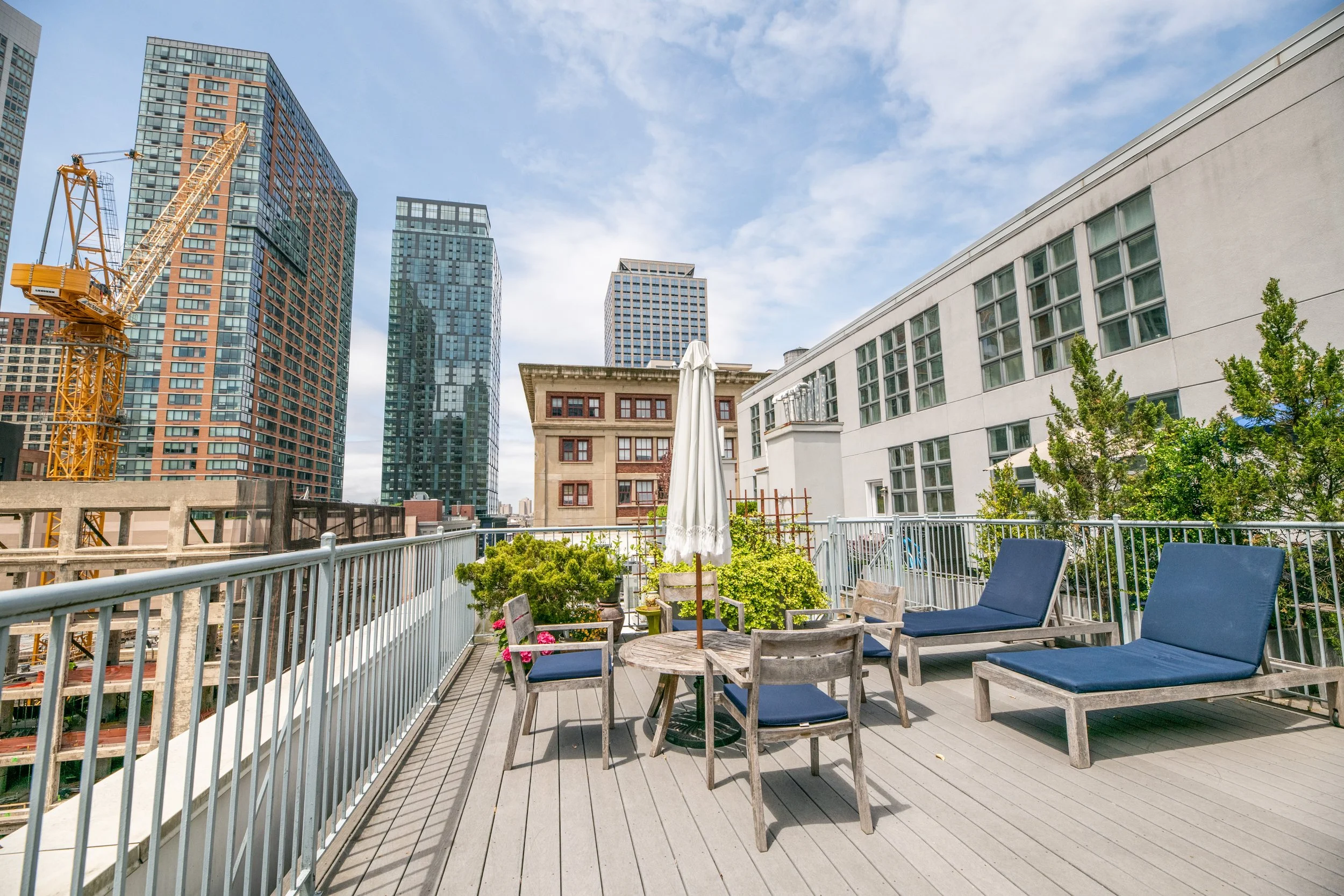 Urban rooftop patio with wooden furniture, lounge chairs, and an umbrella, surrounded by modern high-rise buildings and partly cloudy sky.