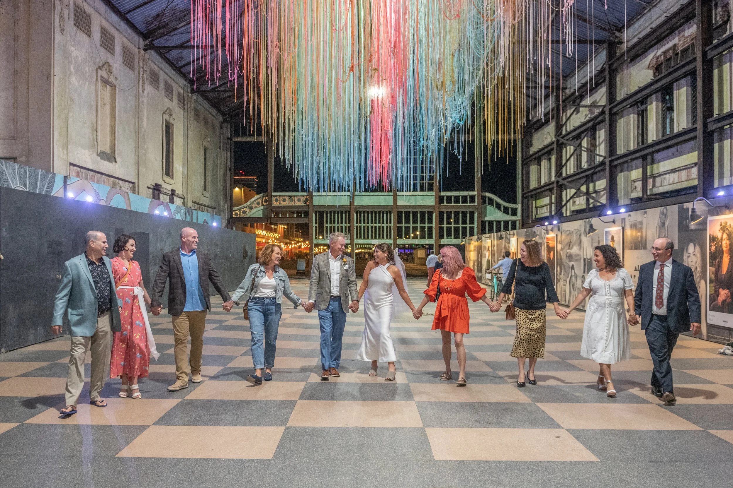 Group of people holding hands and dancing in an indoor space decorated with colorful hanging art installation overhead at night.