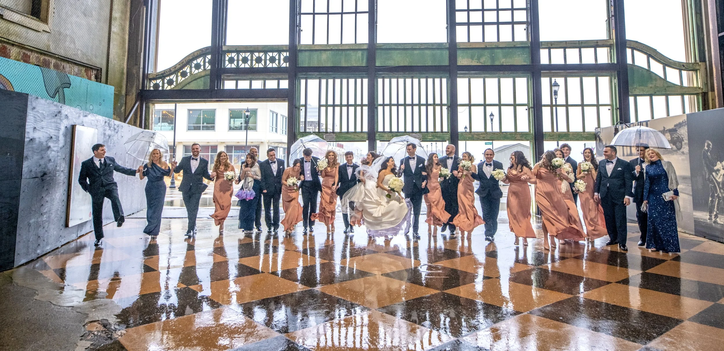Wedding party indoors with rain outside, bride and groom in center, bridesmaids in peach dresses, groomsmen in black tuxedos, people holding umbrellas, checkered tile floor with reflections.