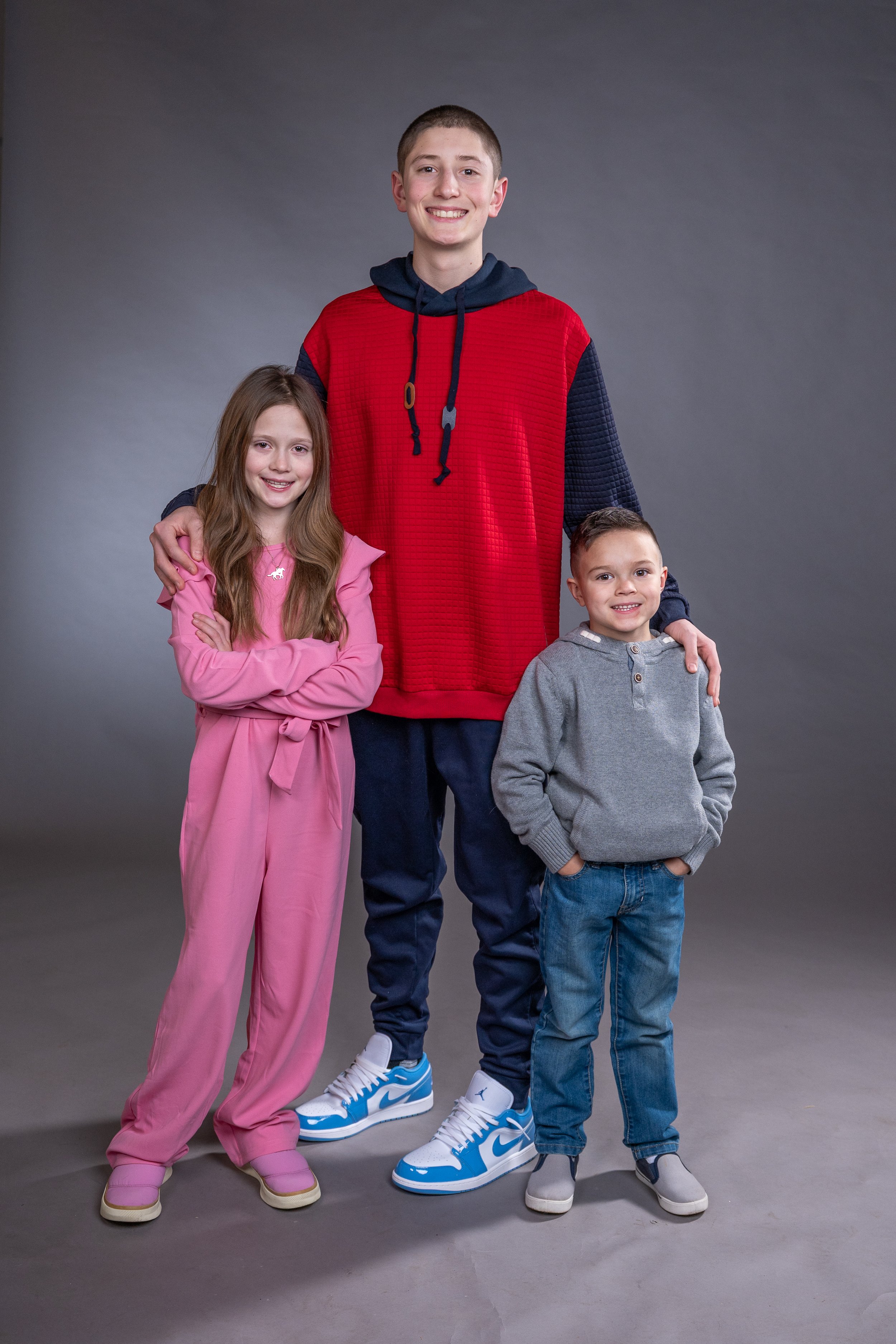 Three kids smiling, standing together against a gray background. The tallest boy is wearing a red and navy hoodie, blue pants, and blue Nike sneakers. The girl in pink jumpsuit has long brown hair, and the boy in gray hoodie and jeans has short dark hair.