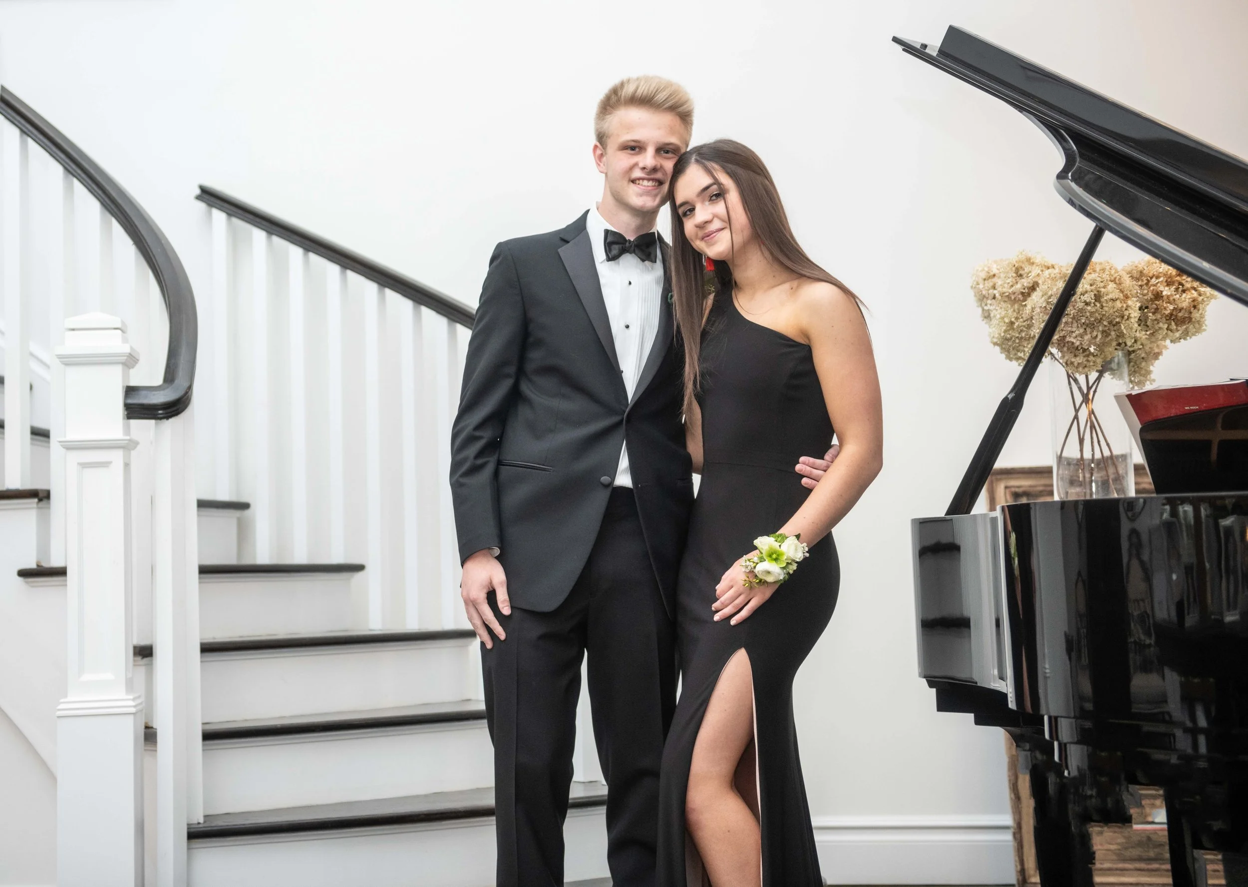 A young man and woman dressed formally, standing side by side, smiling at the camera in front of a staircase with a white wall, a grand piano, and flowers.