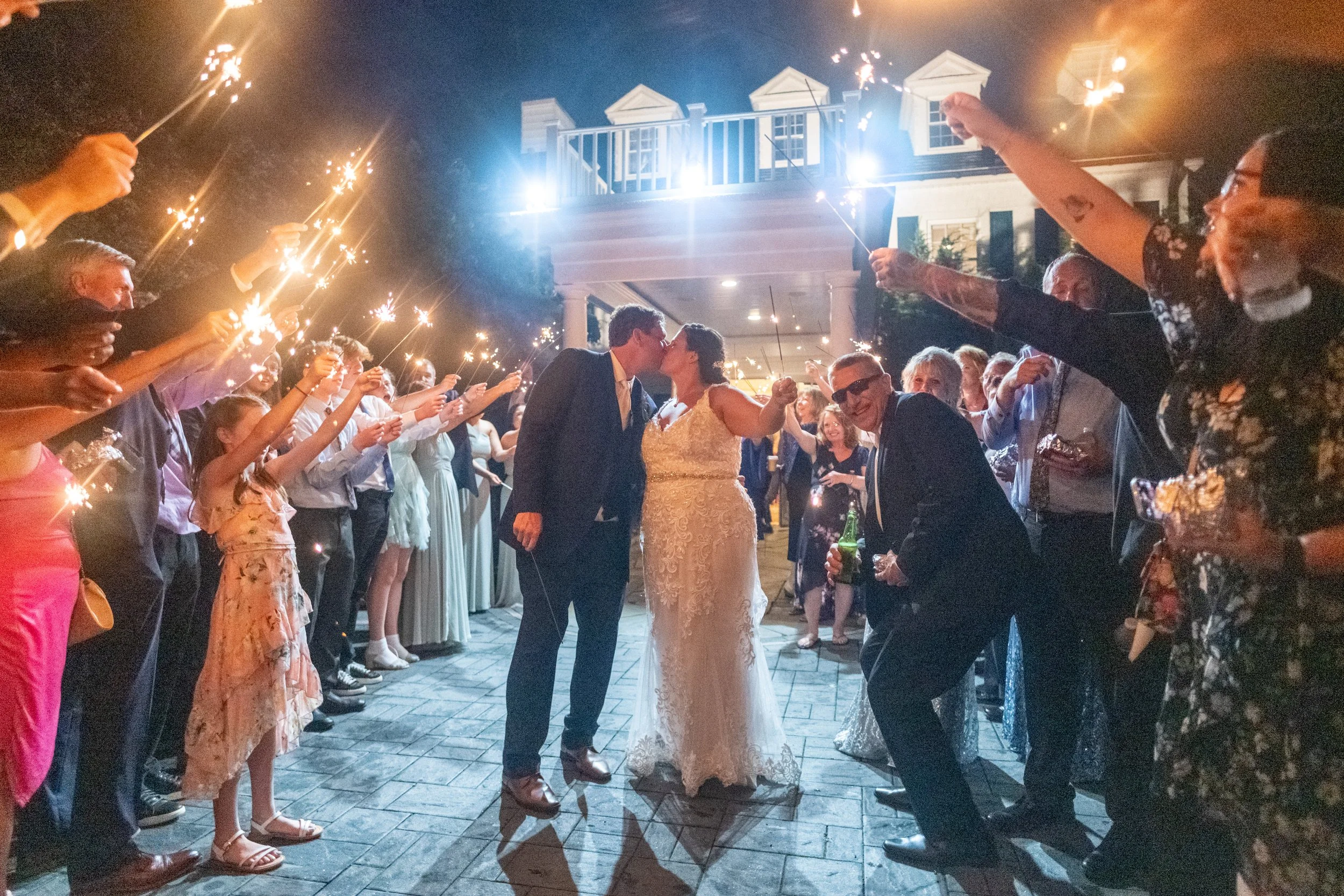 A bride and groom kiss in the center of a celebration surrounded by guests holding sparklers, outside a house at night.