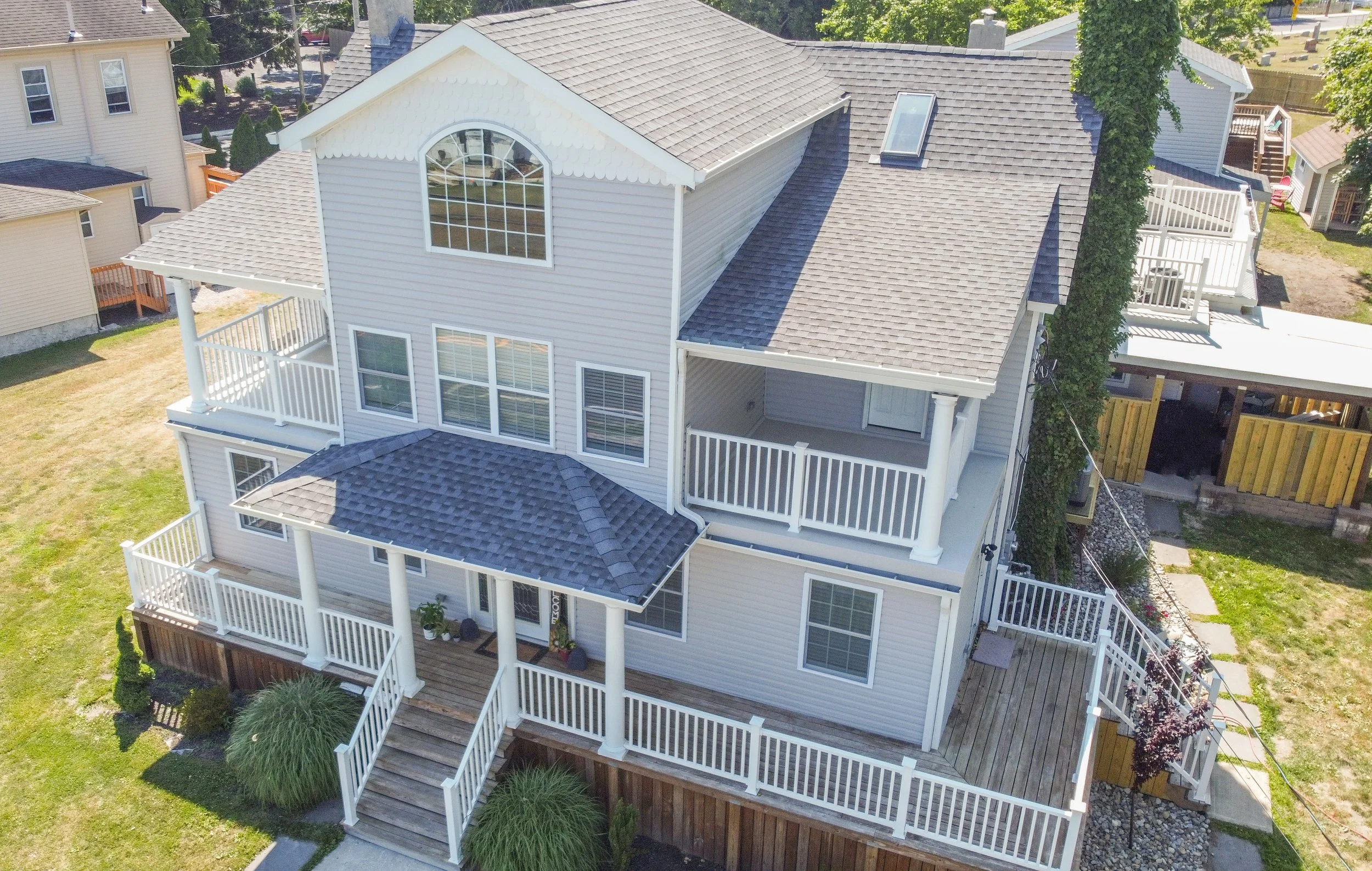 A three-story white house with multiple decks and a gray shingled roof, surrounded by other houses and green lawns.