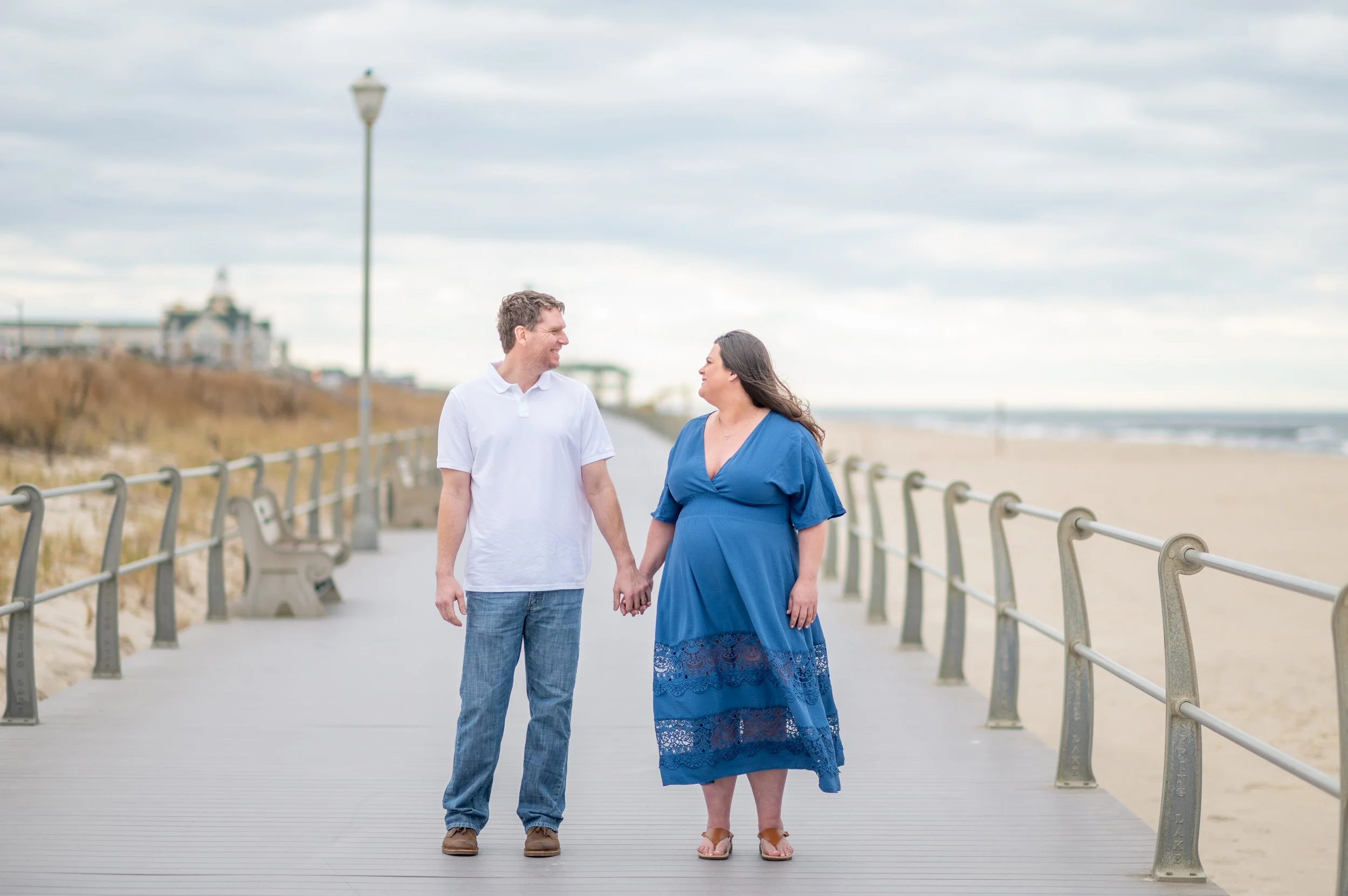 A couple holding hands and walking along a beach pier, smiling at each other in a casual outdoor setting with sand, the ocean, and a cloudy sky in the background.