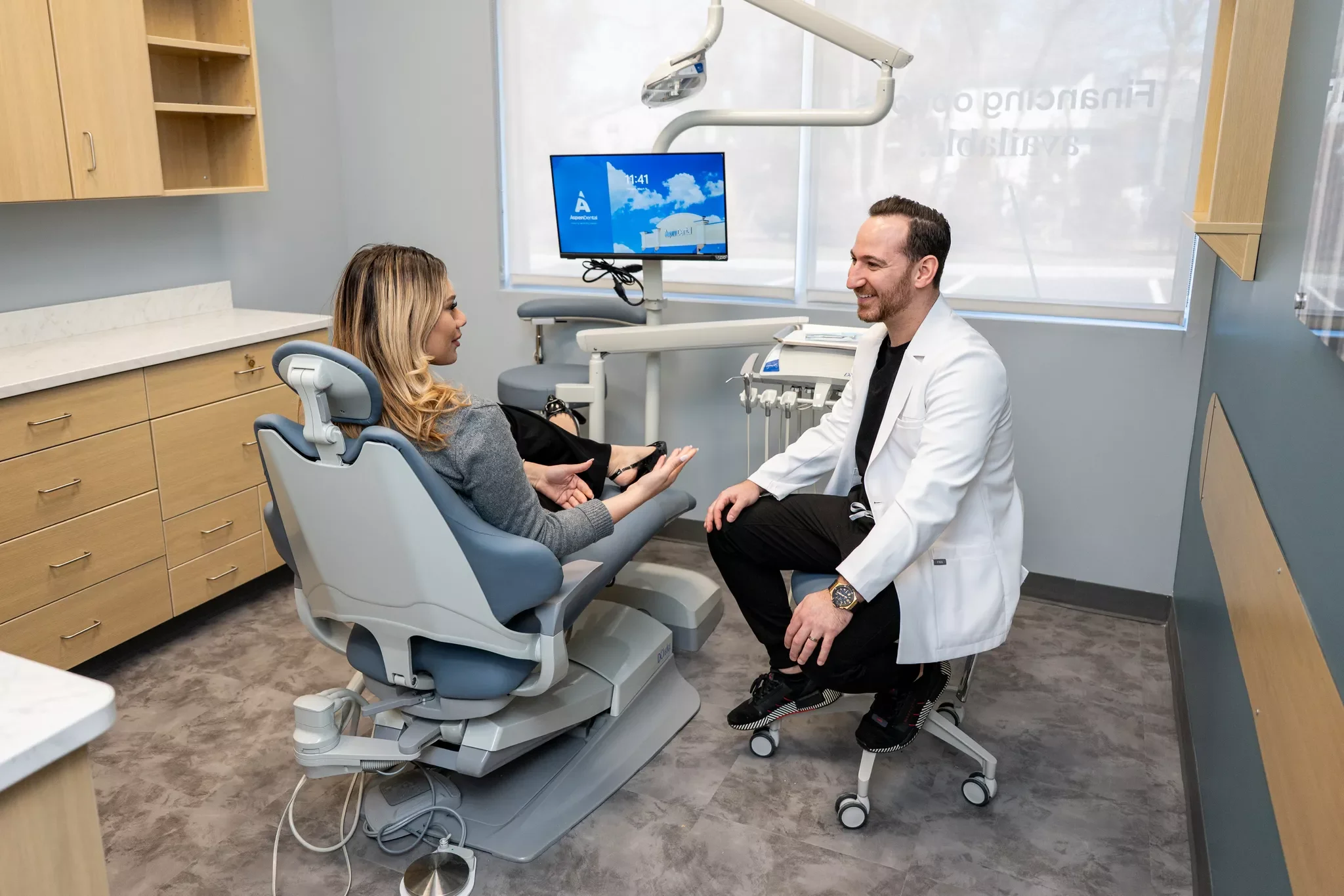 A woman sitting in a dental chair talking to a male dentist who is smiling, in a dental office with medical equipment and cabinets.
