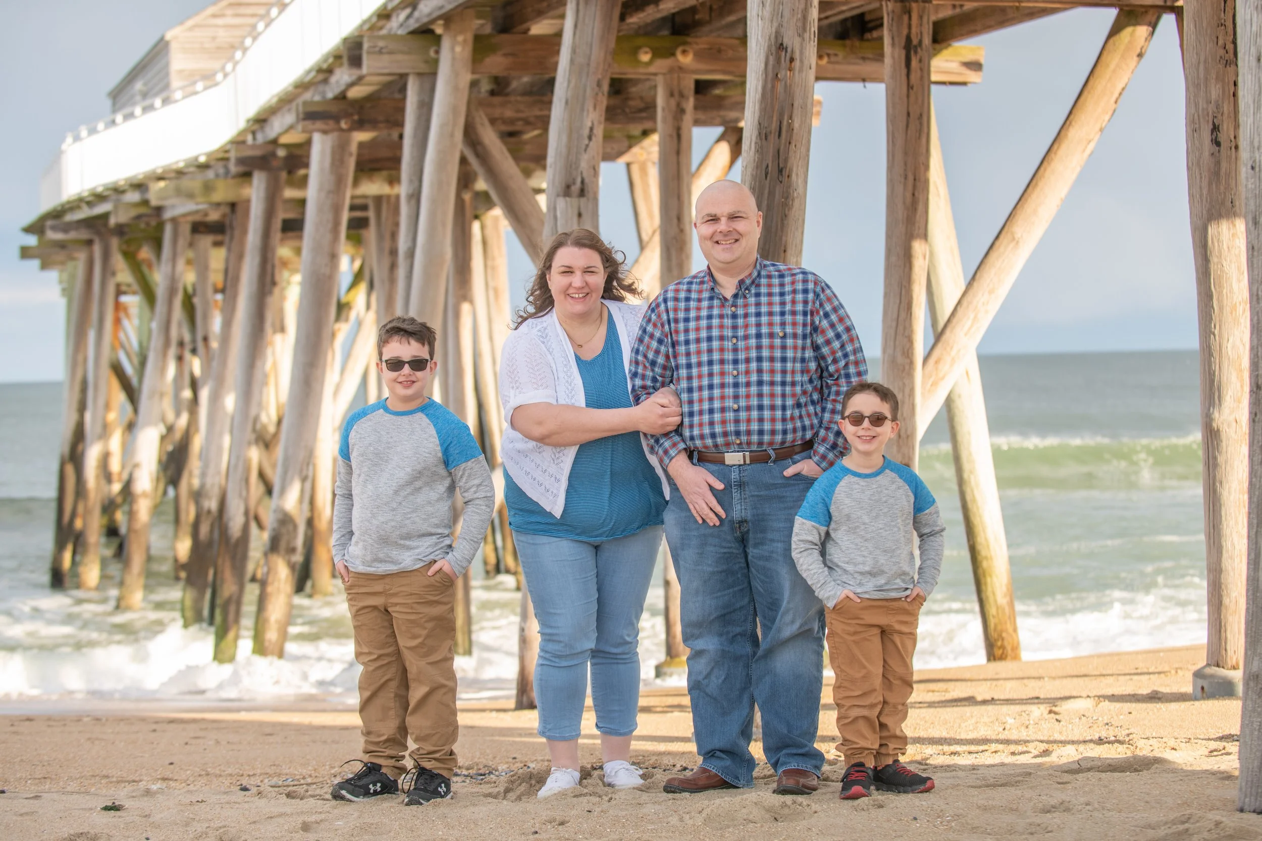 A family of four standing on the beach under a wooden pier, smiling at the camera.