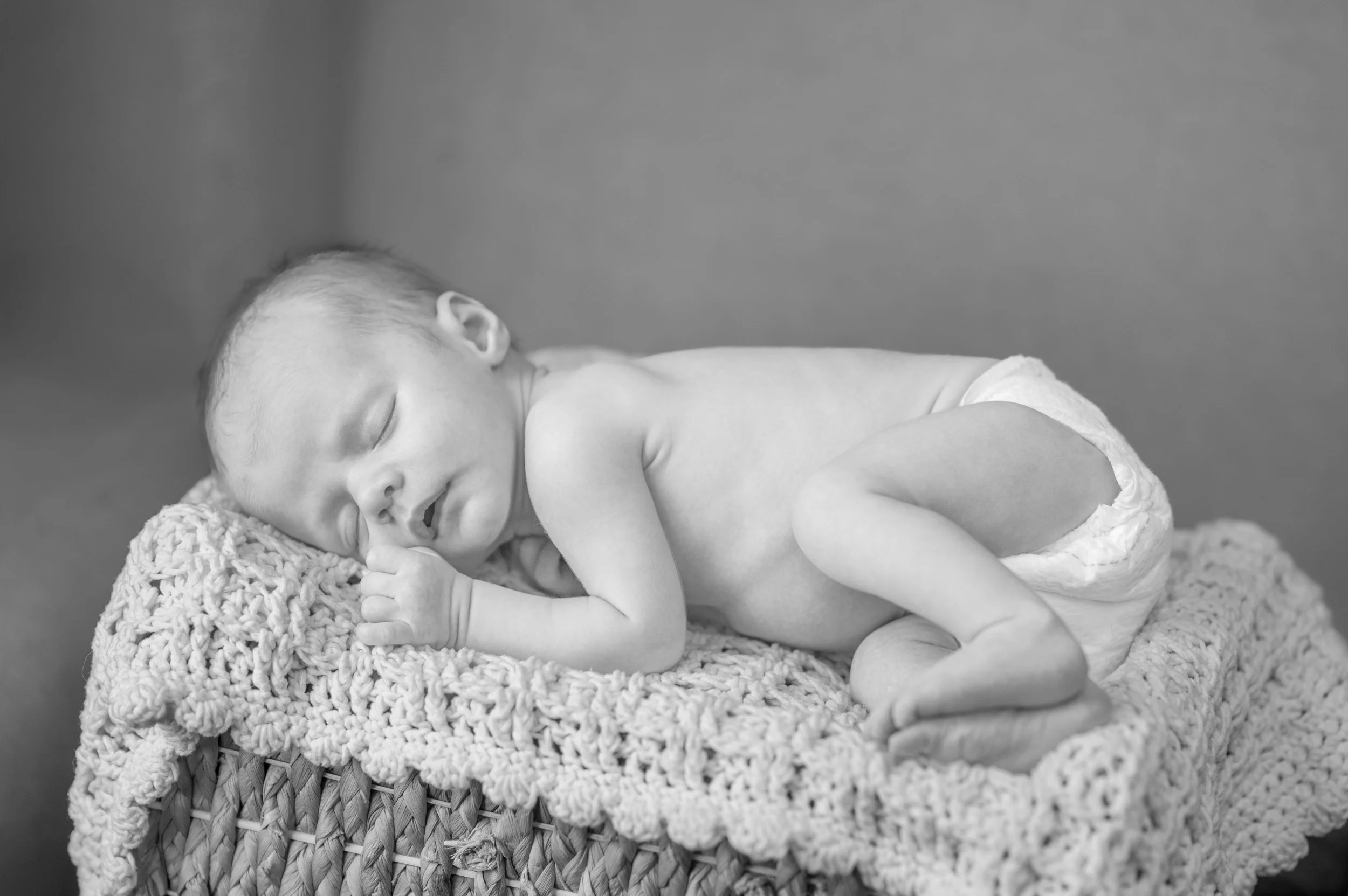 Black and white photo of a sleeping newborn baby lying on a textured blanket in a woven basket.