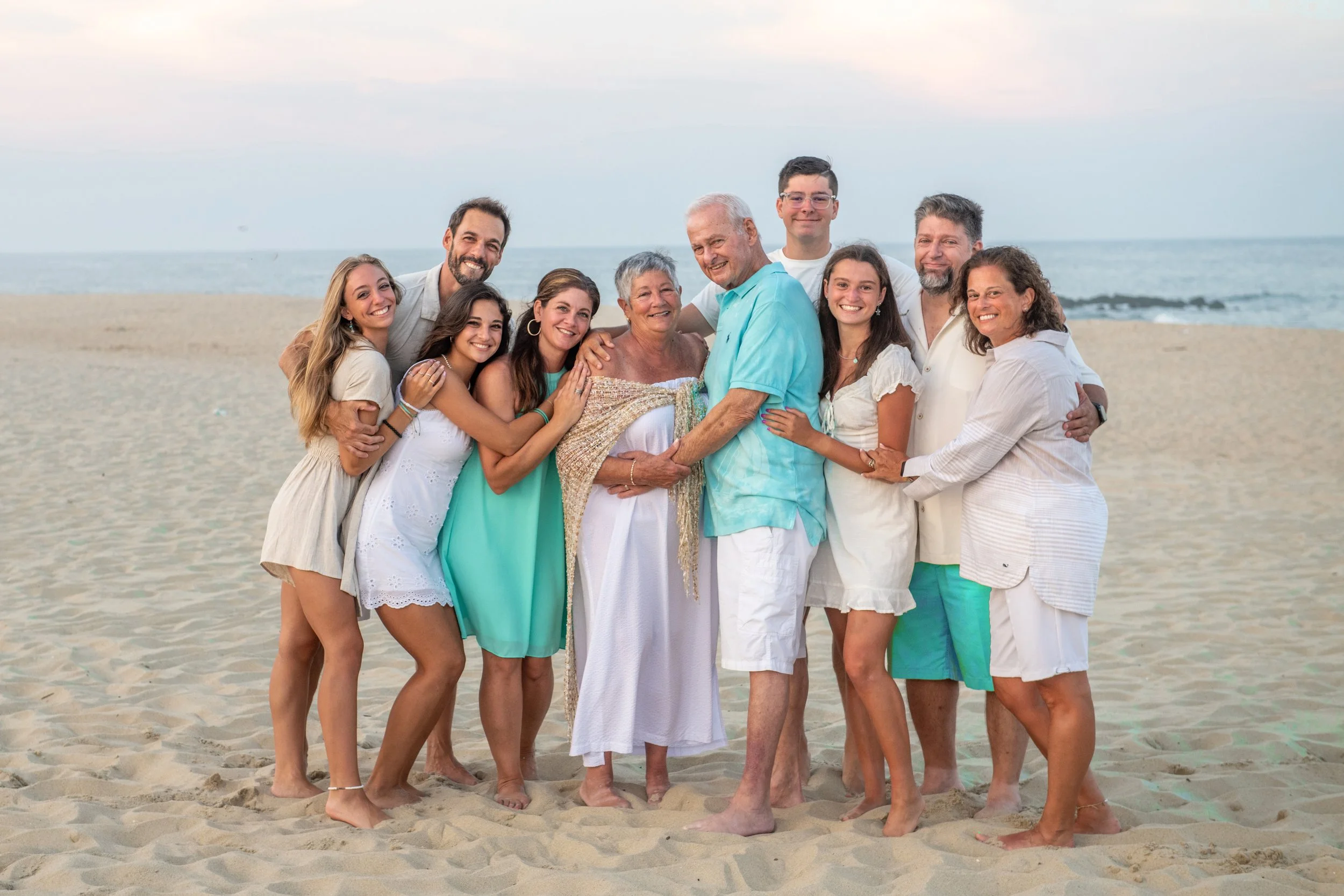 Group of people hugging and smiling on a beach, with the ocean and sky in the background.