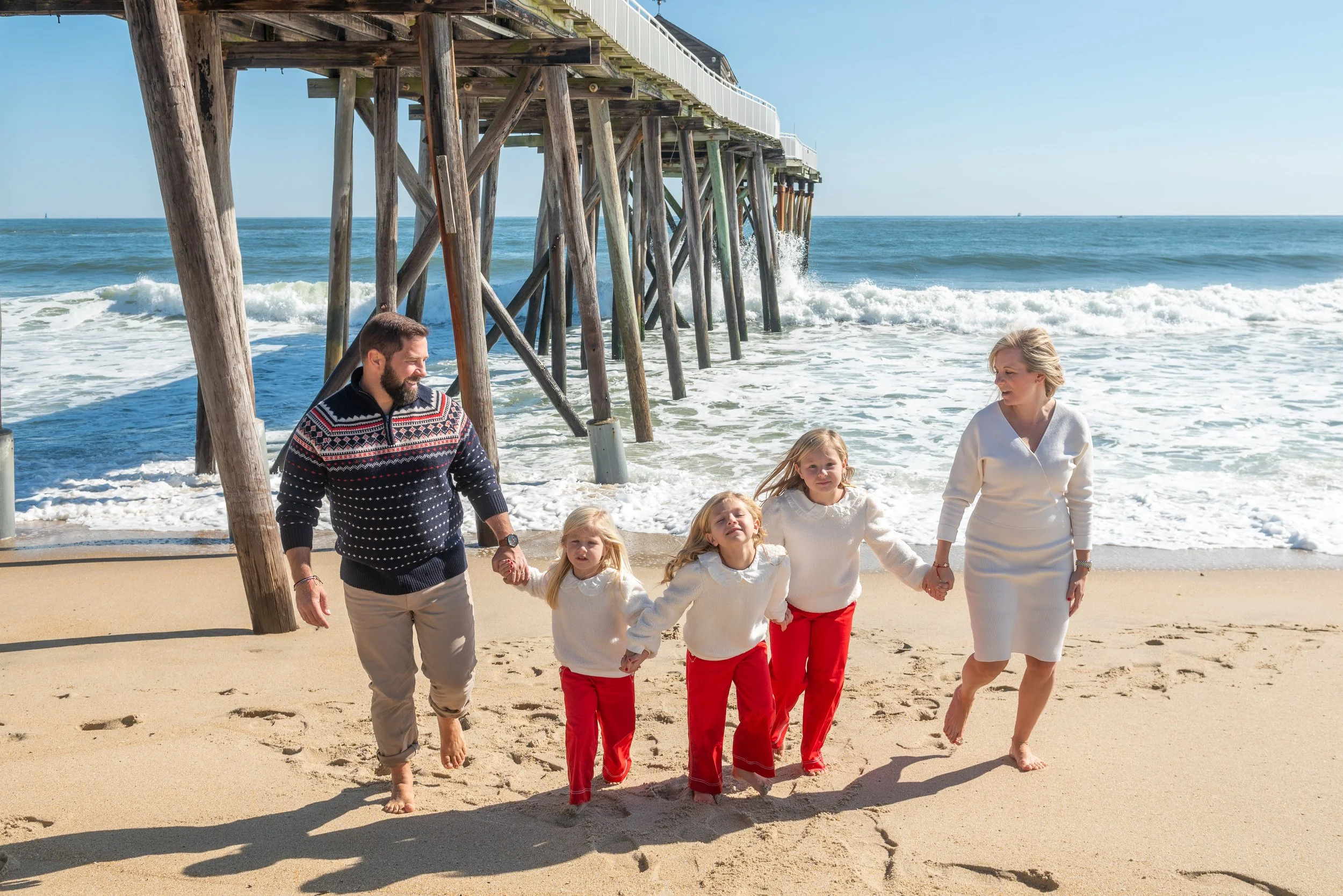 A family of six with three young girls and two adults walk barefoot on a sandy beach near a pier, holding hands, with ocean waves in the background on a sunny day.