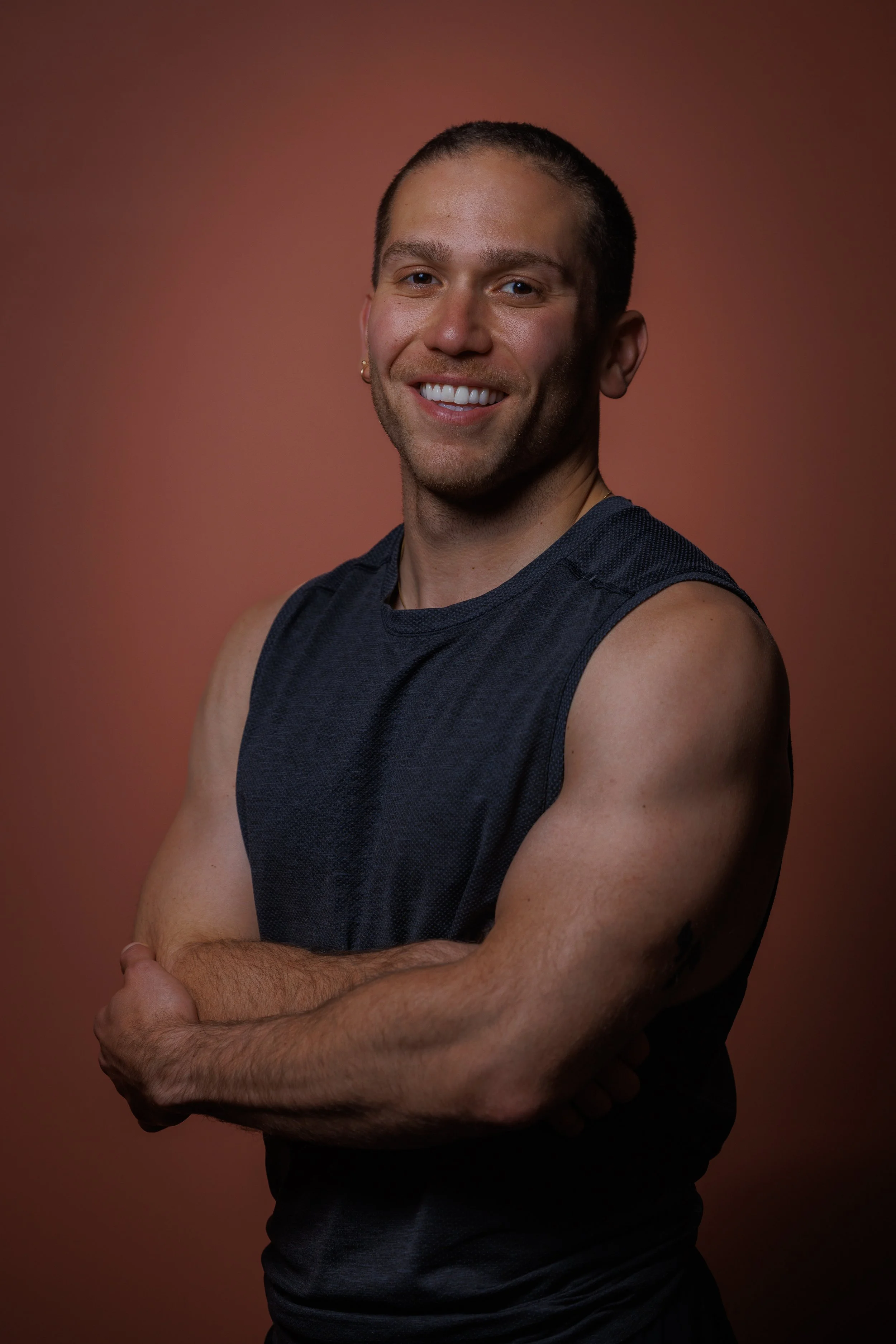 A smiling man with short hair, wearing a sleeveless dark athletic shirt, standing against a plain, reddish-brown background with arms crossed.