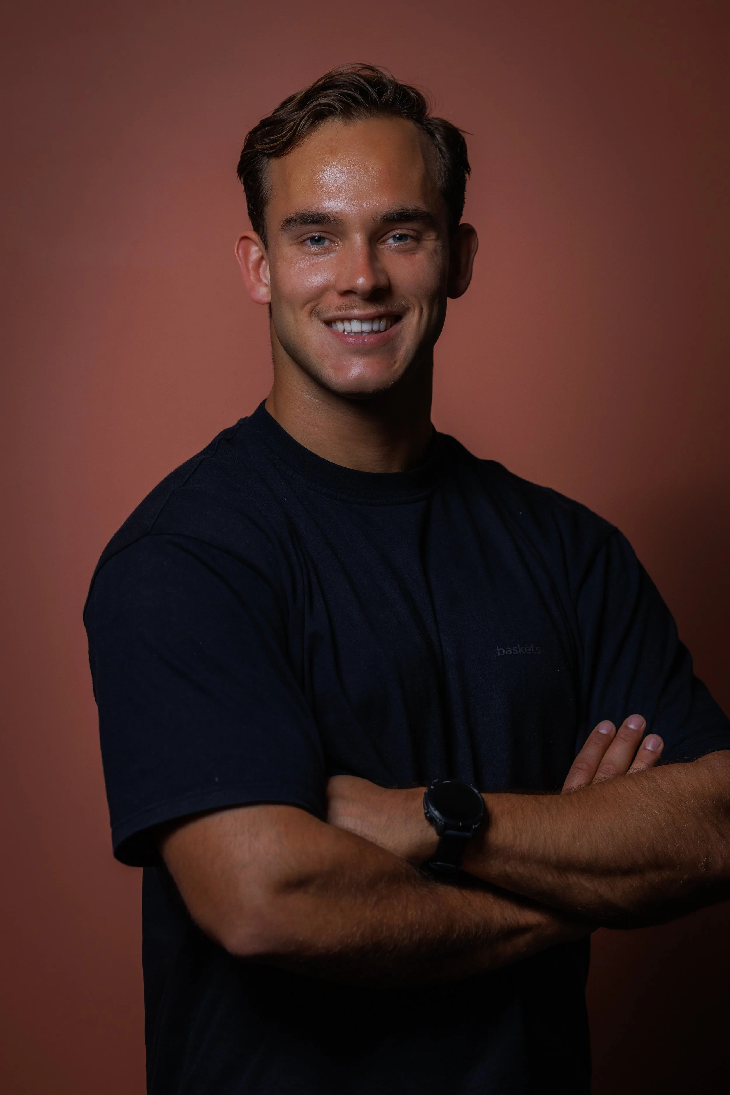 Portrait of a young man with short brown hair, blue eyes, smiling, crossed arms, wearing a black t-shirt and a black wristwatch, against a reddish-brown background.