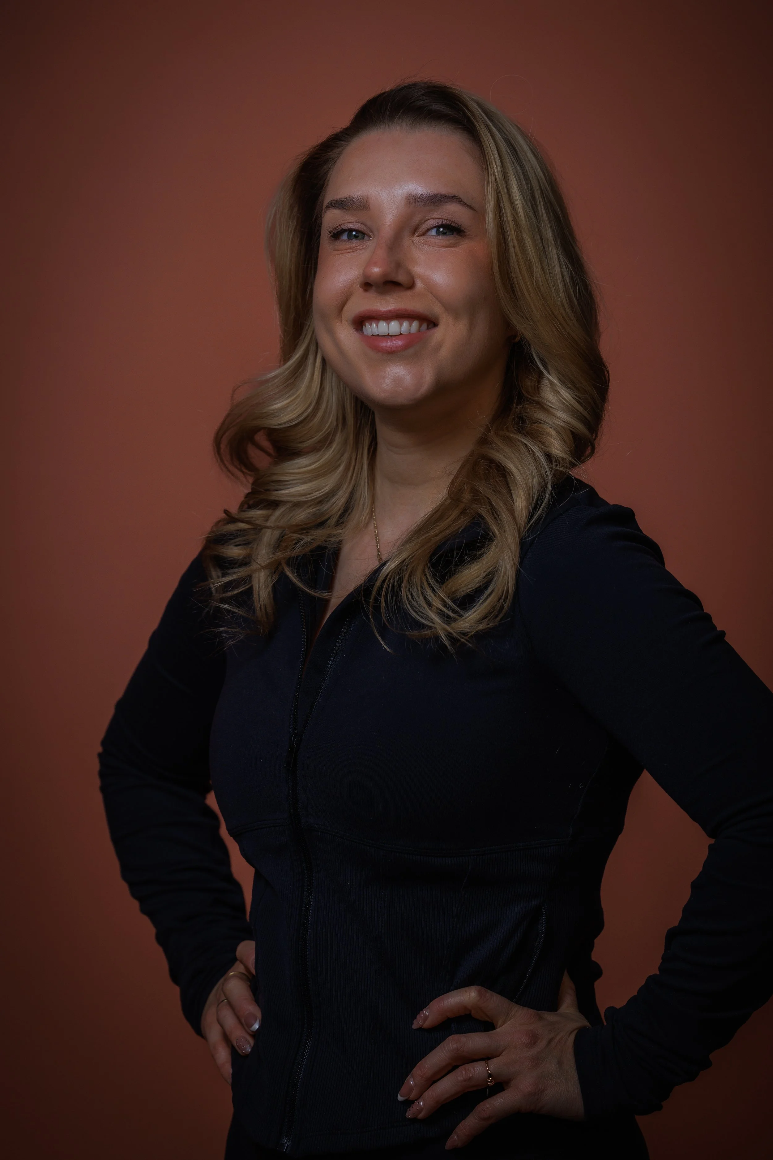 A woman with blonde, wavy hair and light skin, wearing a dark zip-up top, standing with hands on hips against a brownish background, smiling at the camera.
