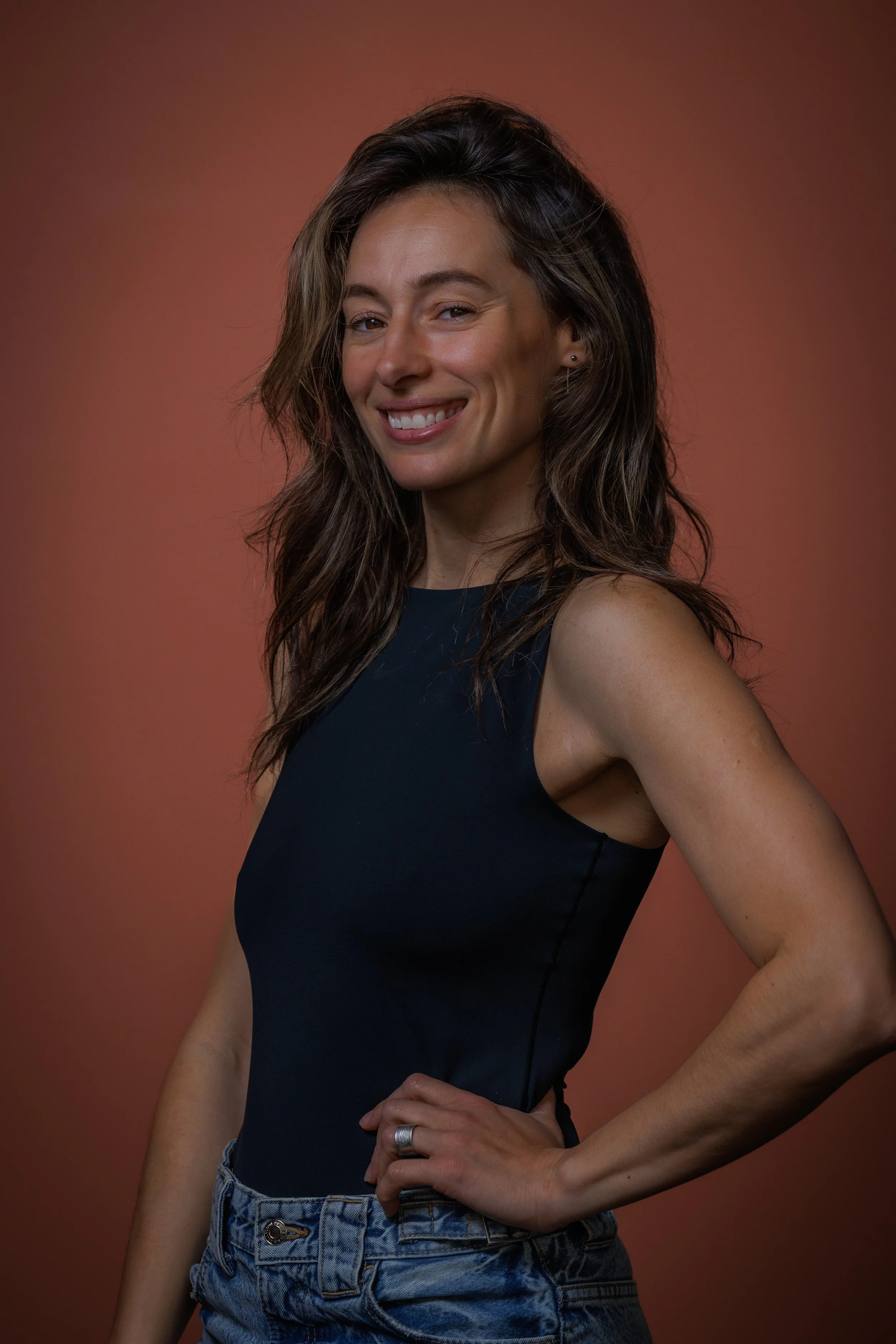 A woman with long wavy brown hair, wearing a sleeveless navy top and high-waisted denim shorts, stands against a plain background. She has a slight smile, one hand on her hip, and is looking relaxed and confident.