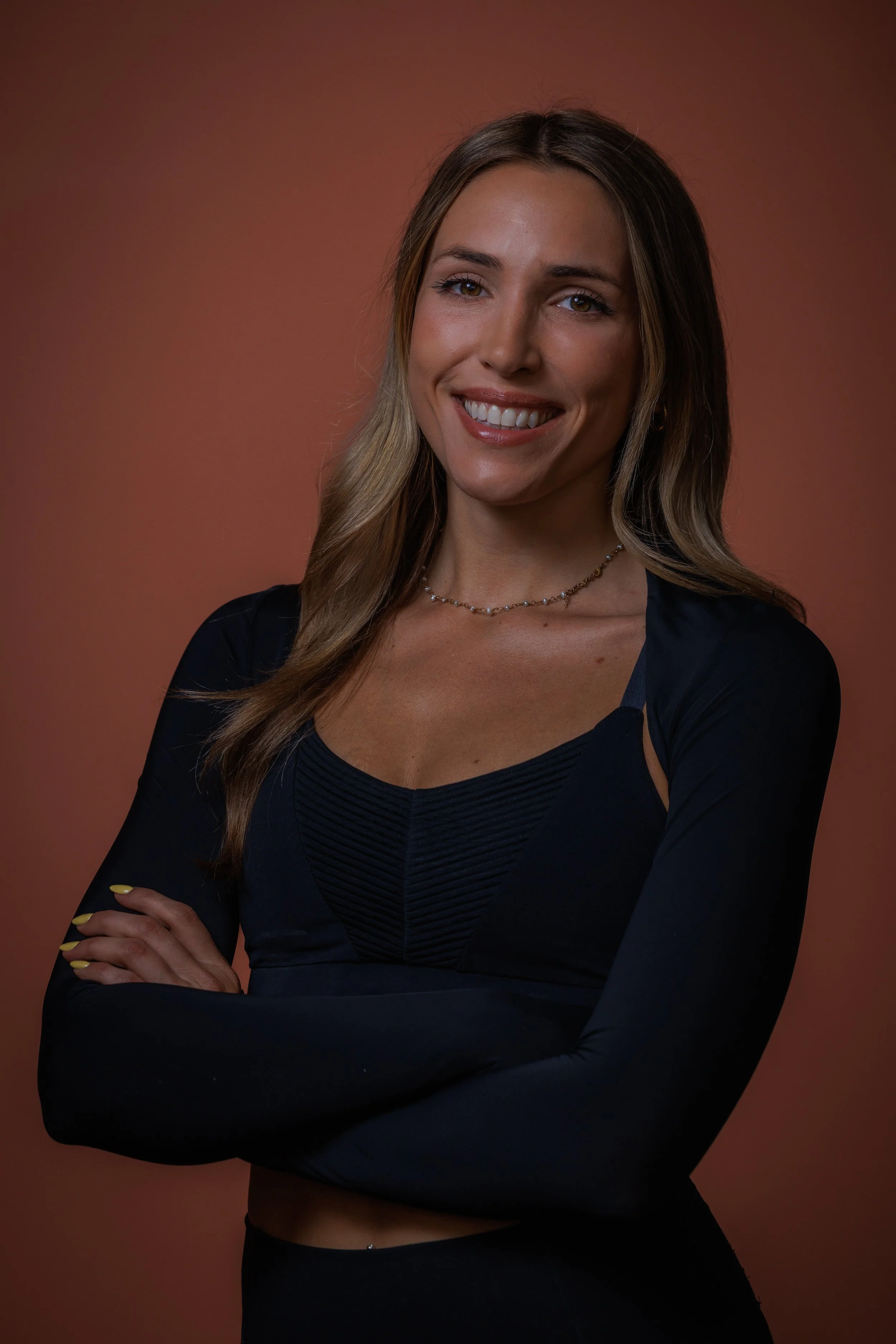A woman with long wavy hair, smiling, wearing a dark long-sleeve top with a v-neck and a beaded necklace, standing against a warm-toned background.