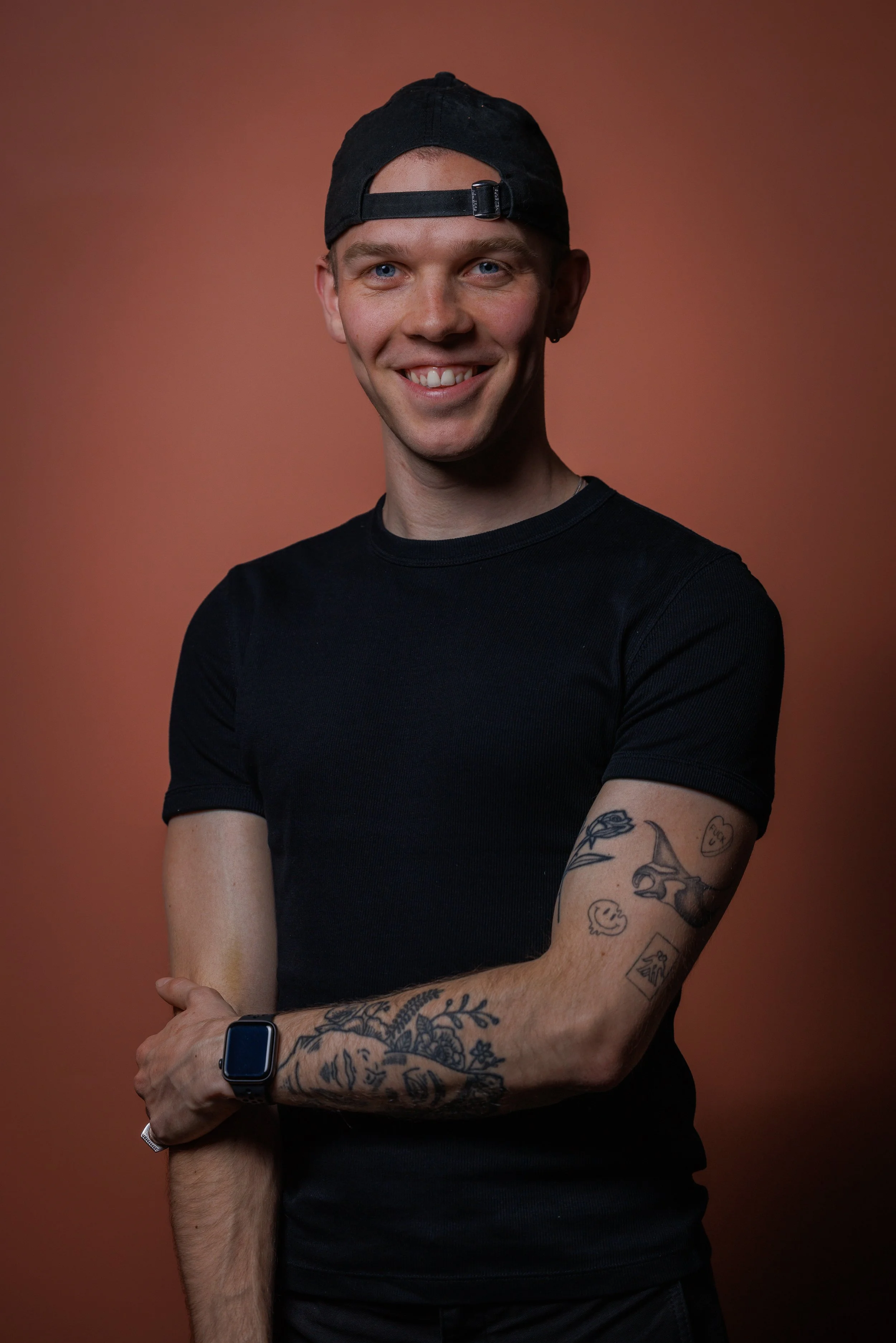 Young man with tattoos on his arms, wearing a black T-shirt, a black baseball cap backward, and a smartwatch, smiling and standing with crossed arms against a plain brown background.