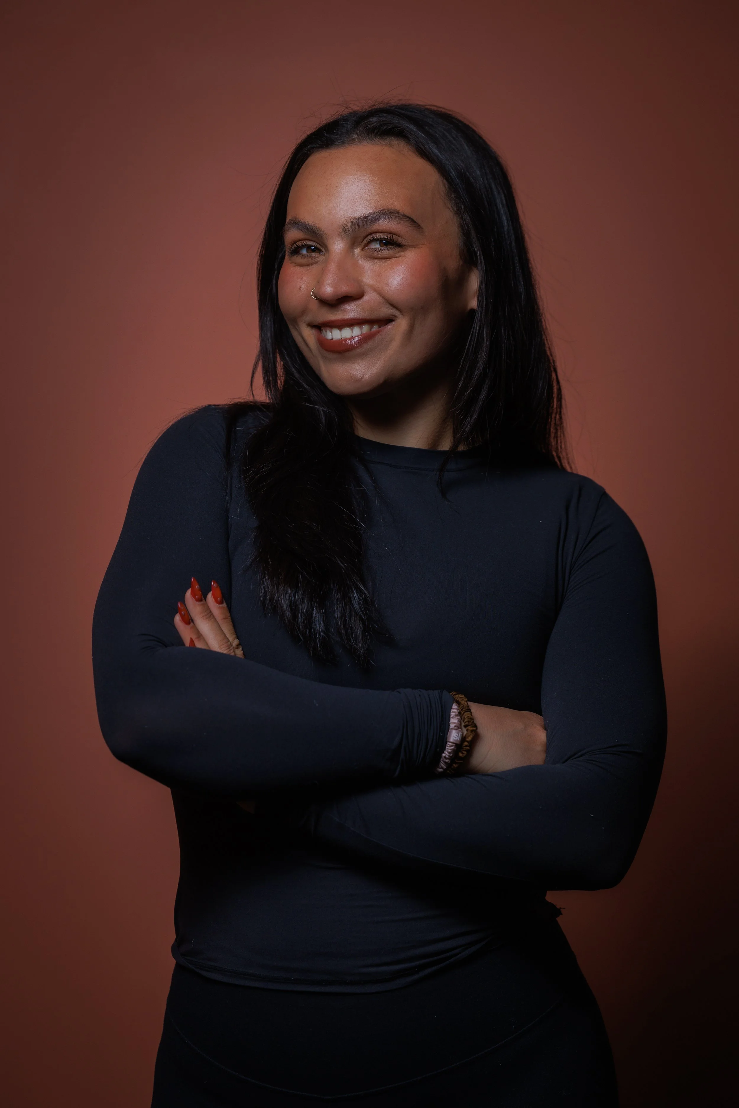 A woman with long dark hair, smiling, wearing a black long-sleeved top, with crossed arms against a brown background.