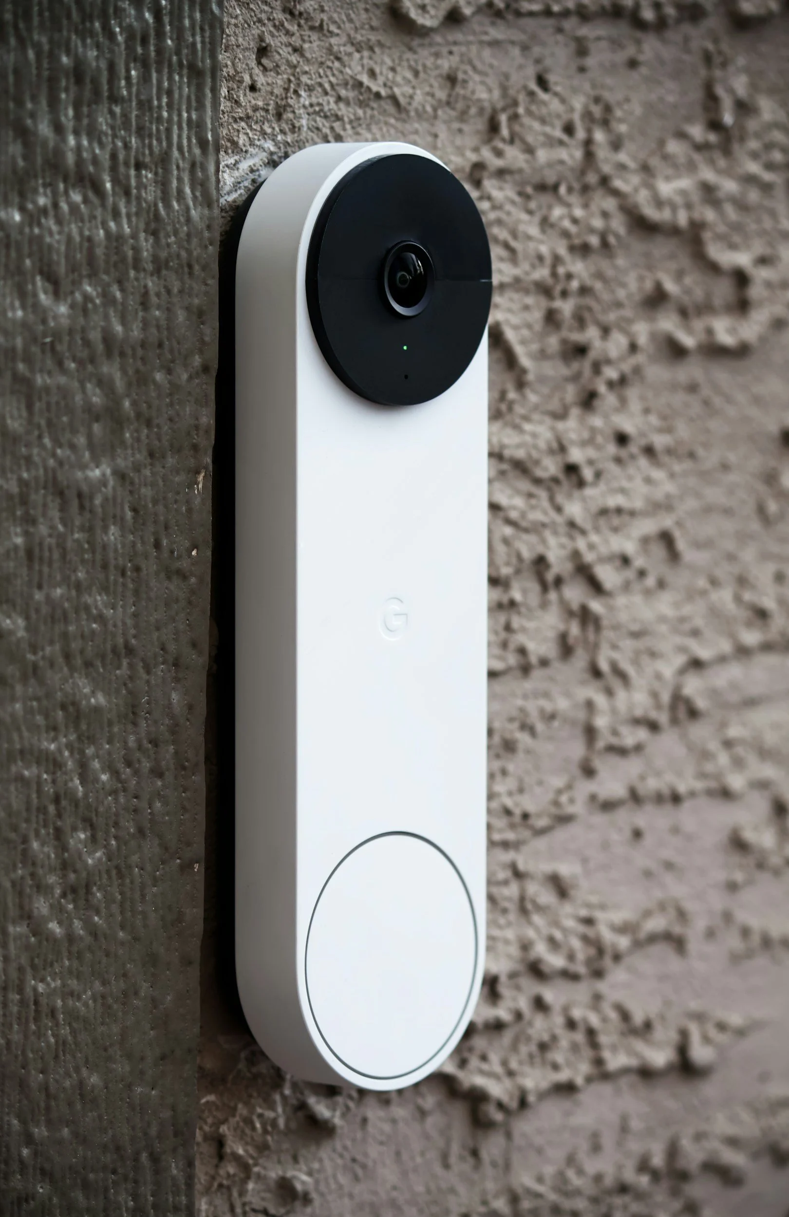 A white video doorbell with a black camera lens mounted on a textured beige brick wall.