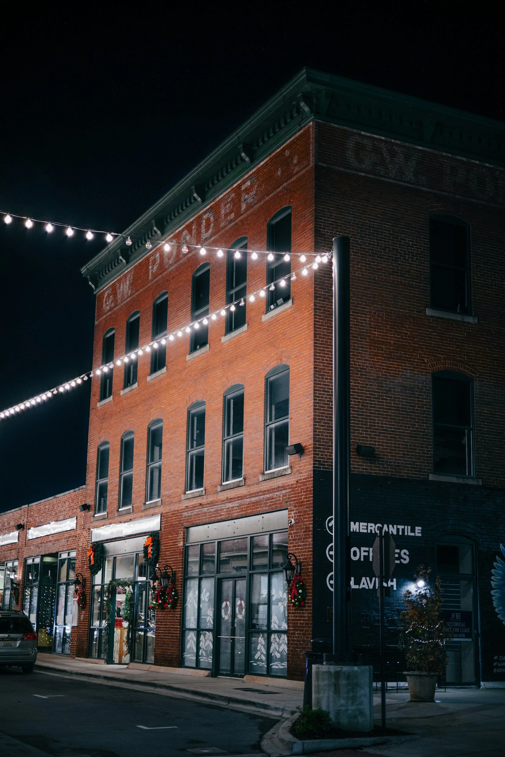 Nighttime street view of a three-story brick building decorated with Christmas wreaths and lights, with a fire department sign faintly visible on the top wall, and string lights hanging across the street.
