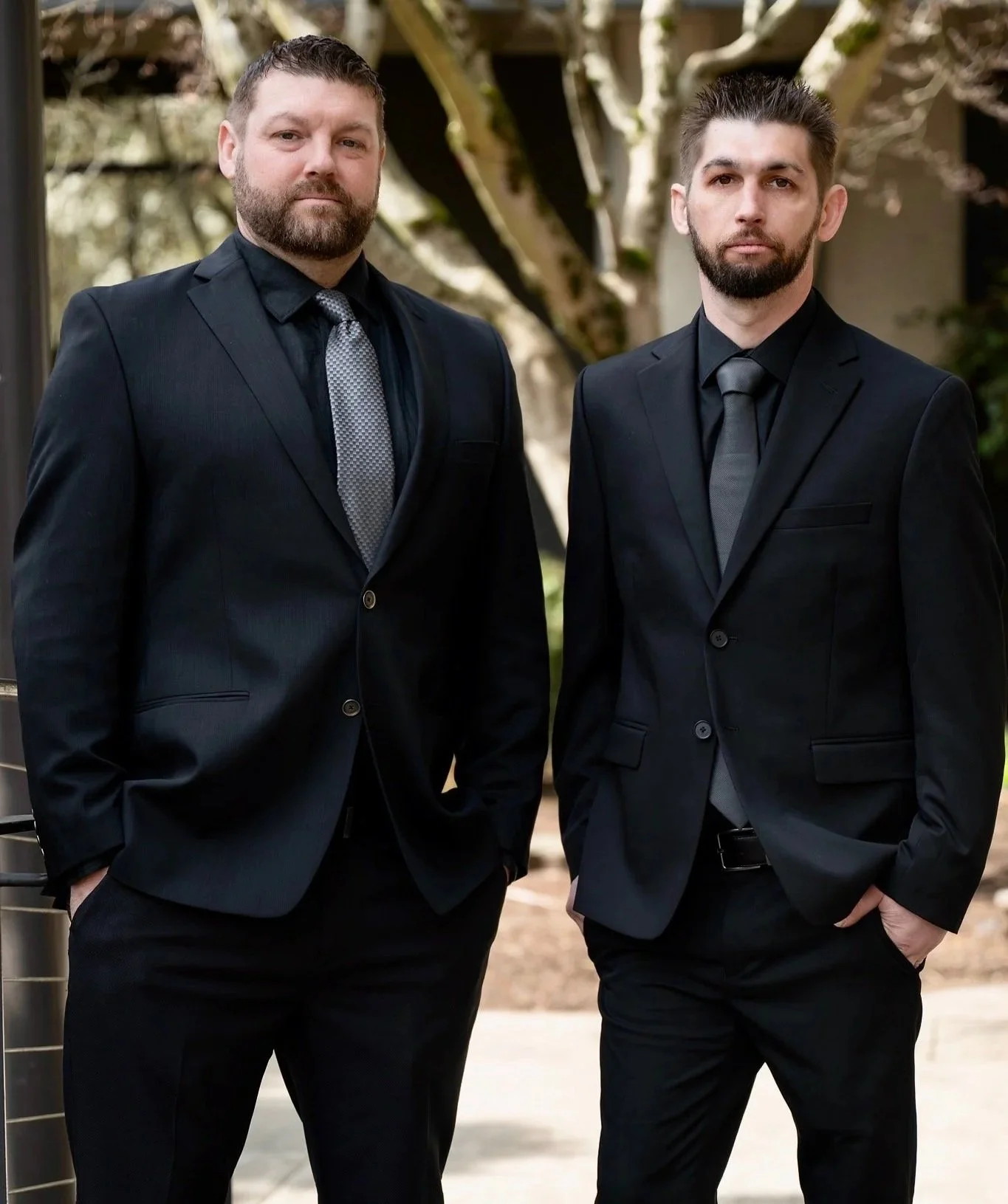 Two men in black suits and gray ties standing outdoors.