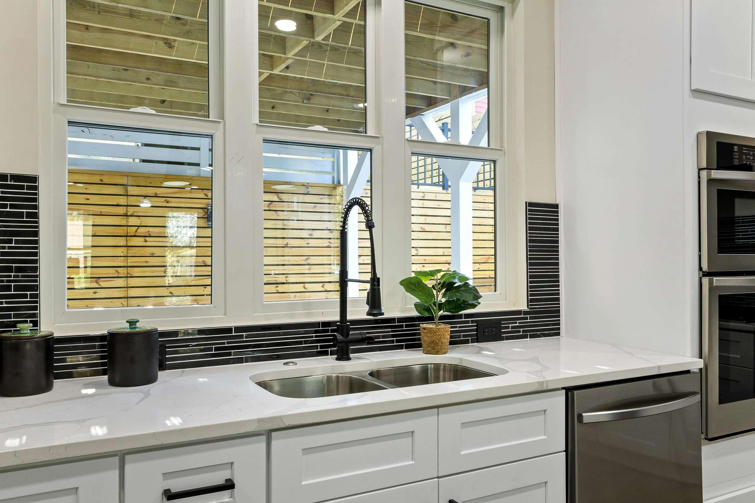 A modern kitchen with a white countertop and a black faucet over a double sink. There is a plant in a woven pot on the counter, and two black ceramic jars. The background features a large window with a view of a wooden fence and an outdoor structure.