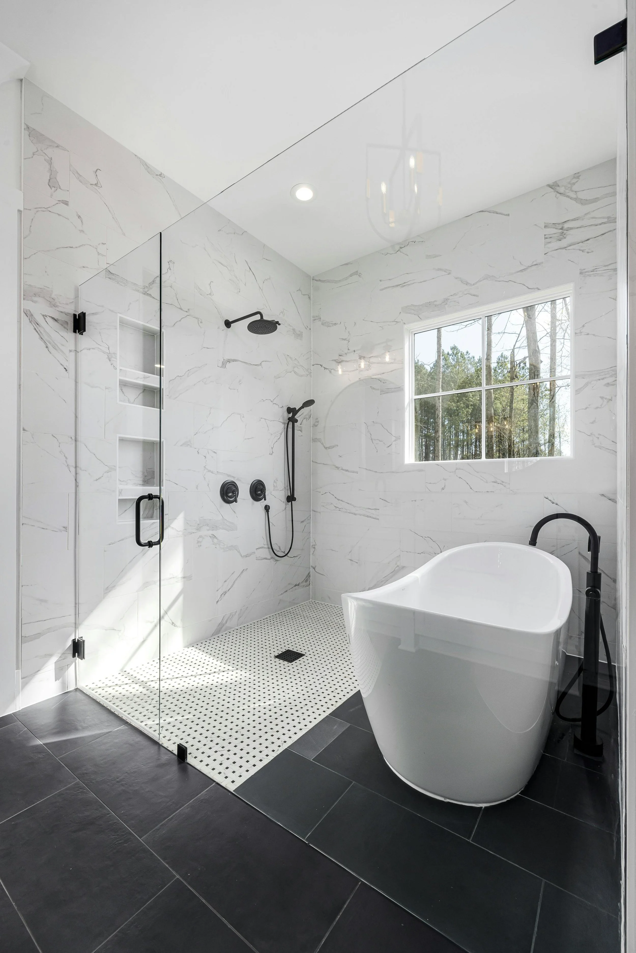 Modern bathroom with a walk-in shower with black fixtures, a mosaic tile floor, a large bathtub, a window showing trees outside, white marble walls, and black floor tiles.
