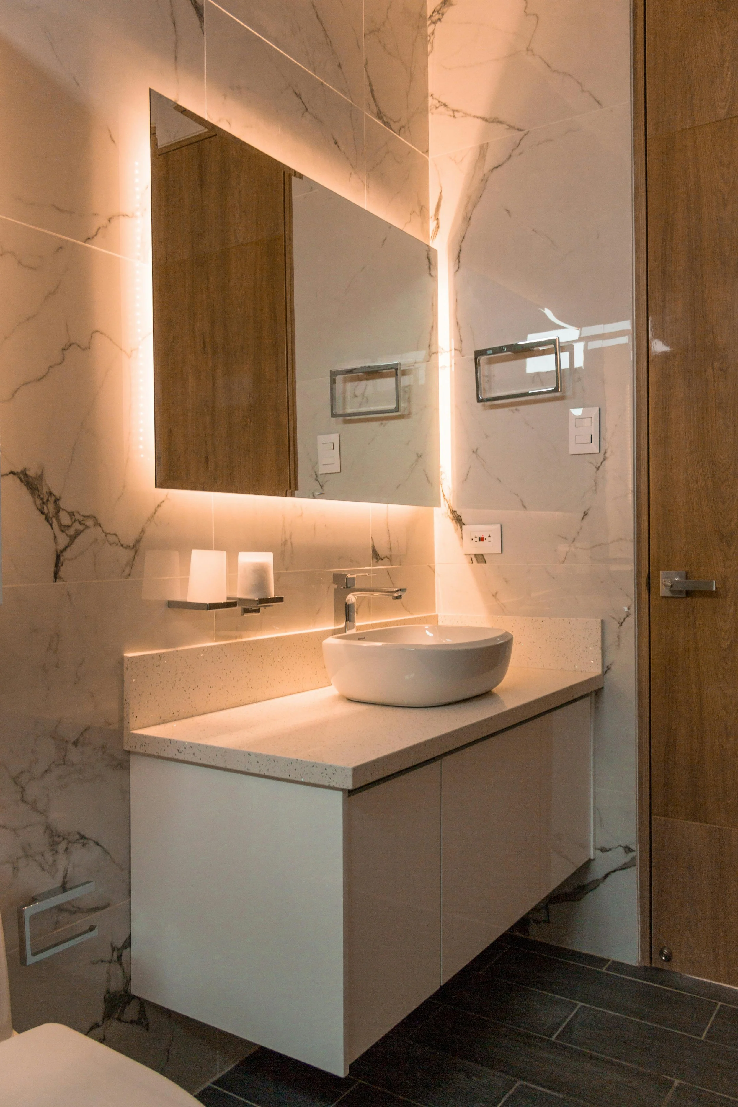 Modern bathroom with marble walls, a white countertop sink, a large mirror with LED lighting, and wooden accents.