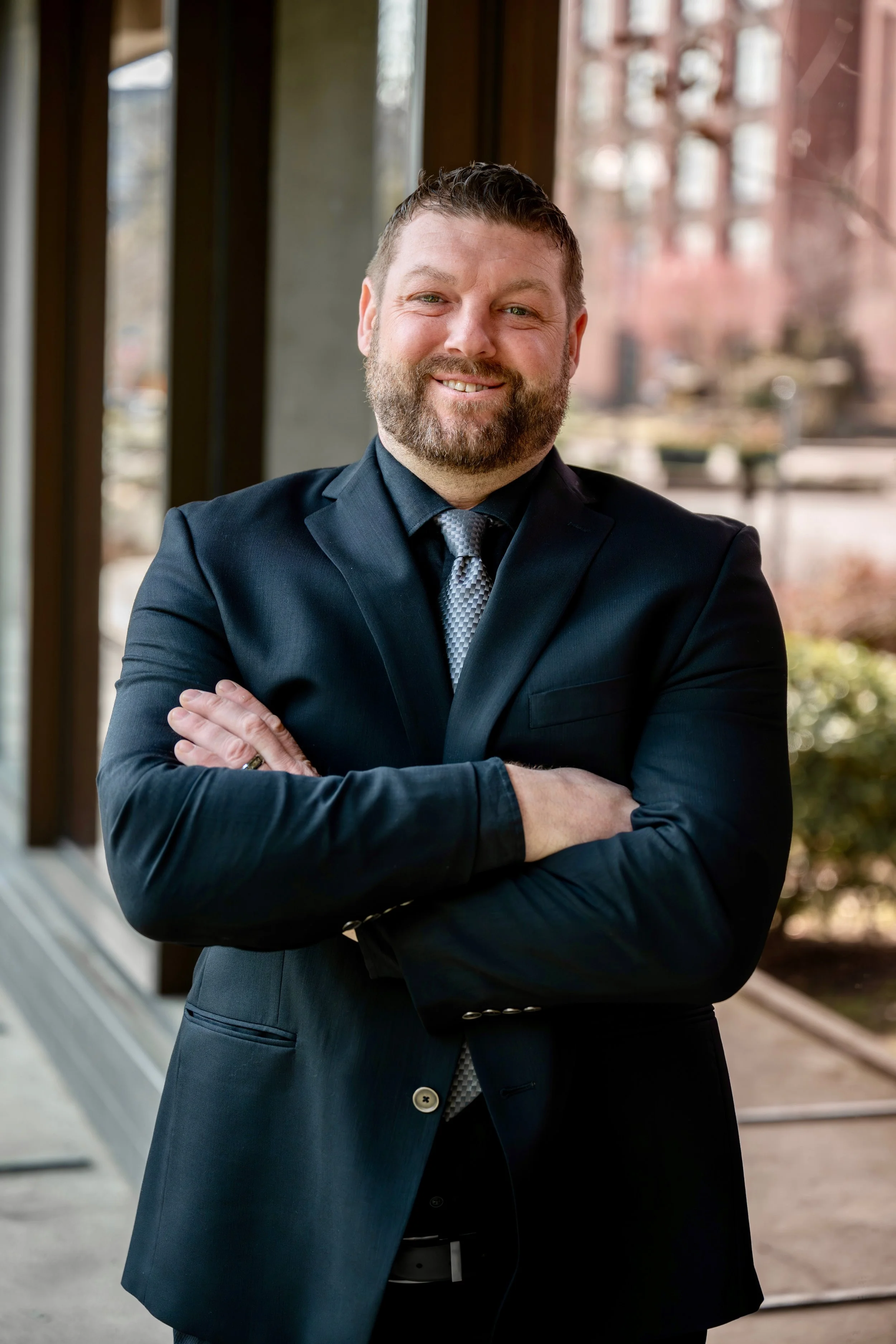 A man with a beard wearing a dark suit and tie, arms crossed, smiling at the camera.