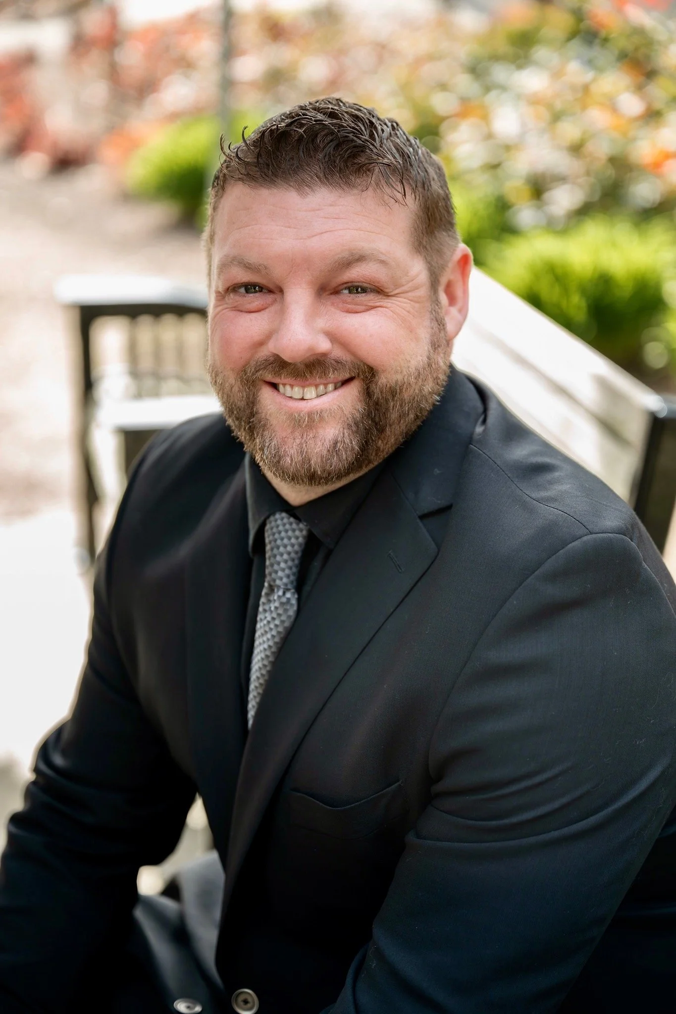 A man with a beard and short hair smiling, wearing a black suit and patterned tie, sitting outdoors near a wooden bench with blurred autumn foliage in the background.