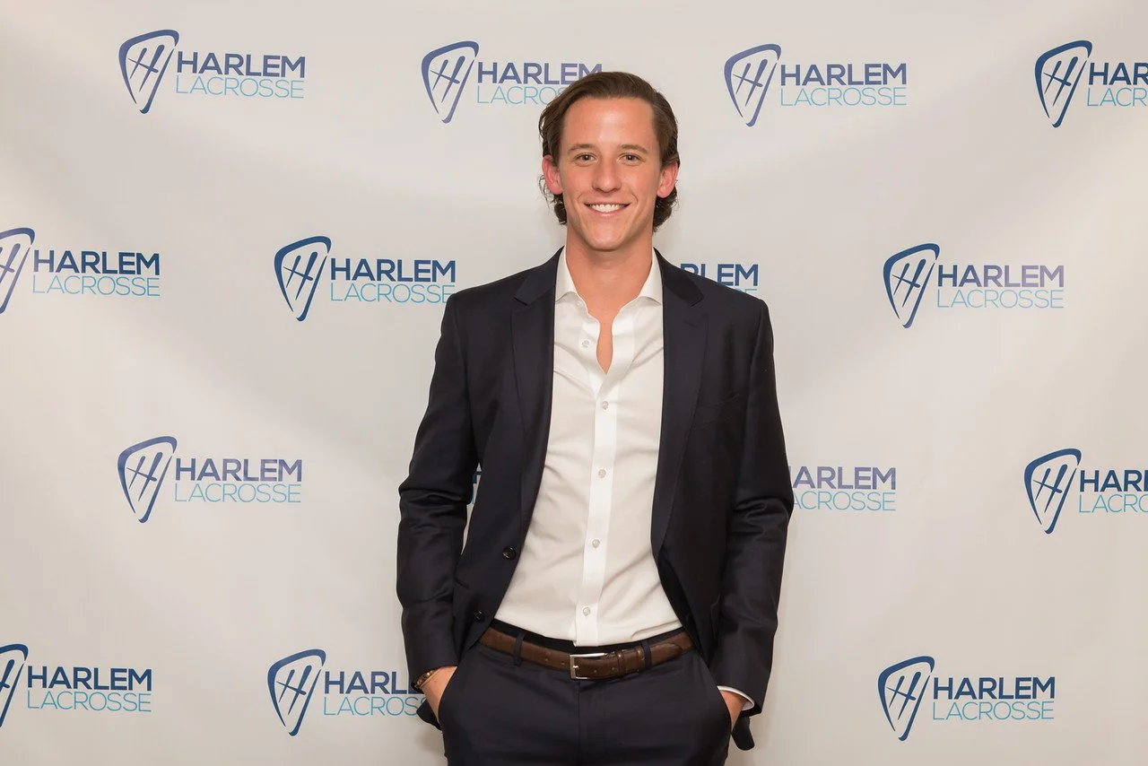 Man in a dark suit and white shirt smiling, standing in front of a Harlem Lacrosse backdrop.