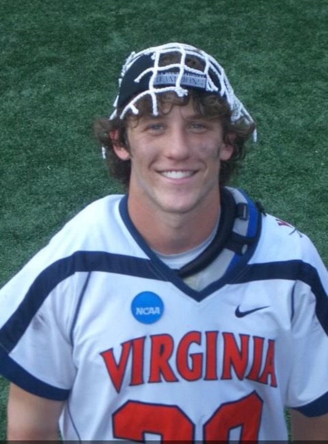 Young man in a Virginia college lacrosse jersey, smiling, wearing a collegiate lacrosse helmet with a protective face mask, on a grassy field.