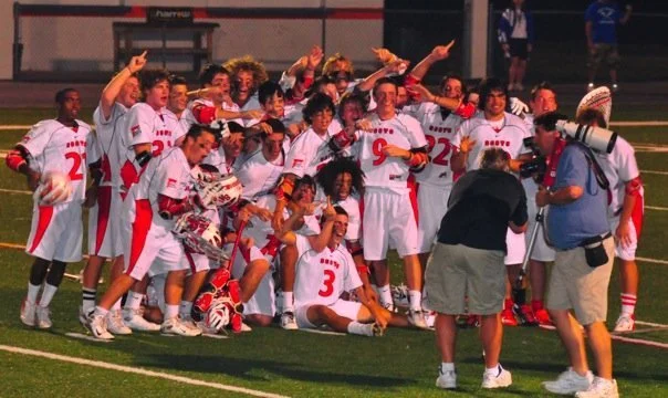 A group of female soccer players in white and red uniforms celebrating with a coach and photographers on a soccer field.