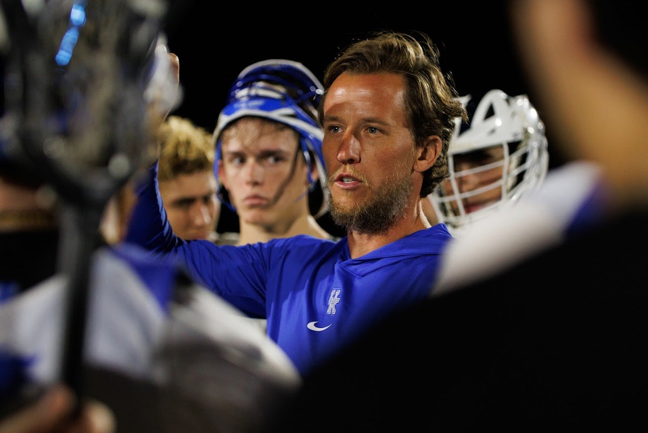A football coach in a blue jacket giving instructions to his team during a timeout, with players listening attentively, some wearing helmets, on a football field at night.