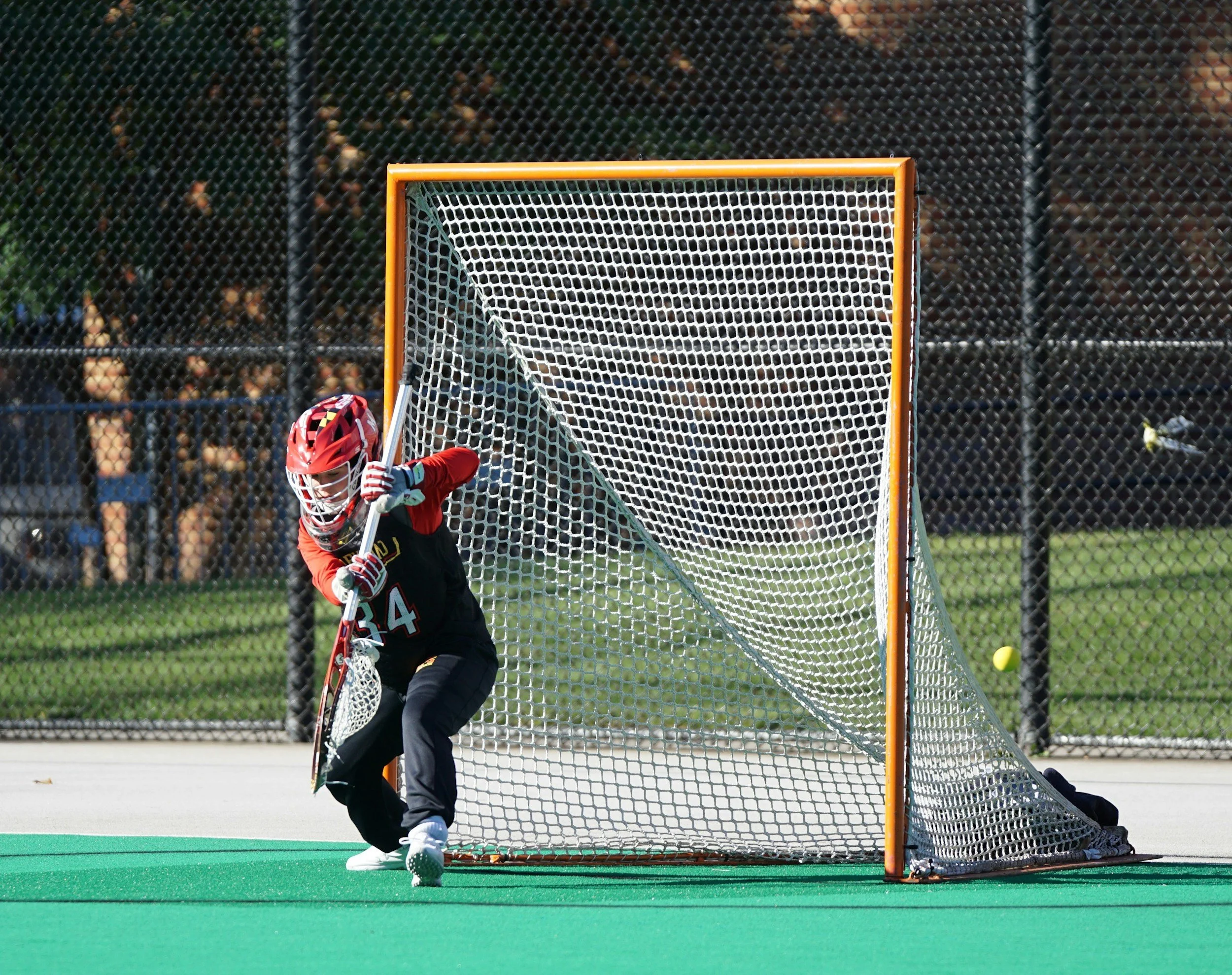 A young field hockey player in a red helmet and uniform attempting to save a ball in front of the goal on an outdoor field.
