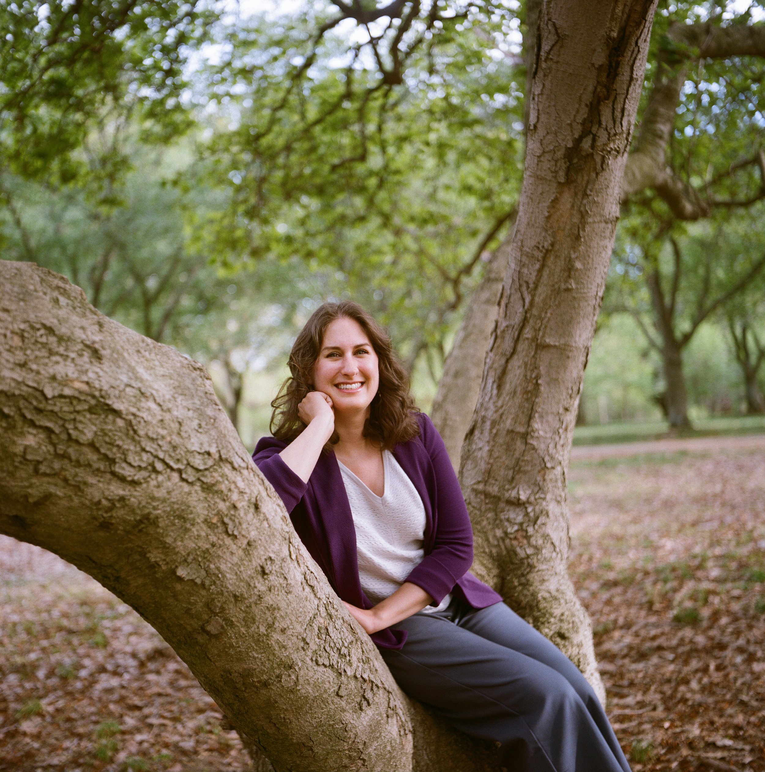 A woman with brown hair smiling and sitting in a tree in a park with green leaves and a walkway in the background.