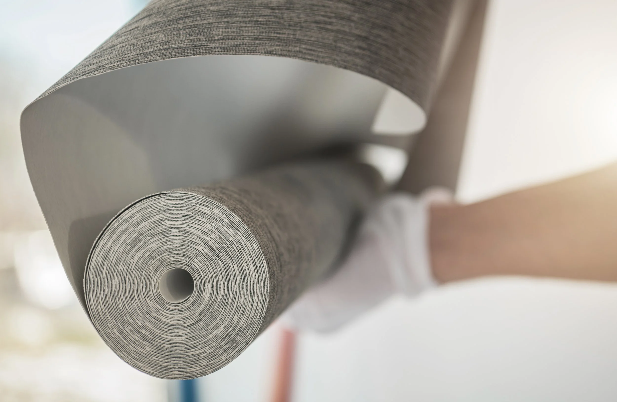 Close-up of a person holding and unrolling a large roll of gray wallpaper or textured paper.
