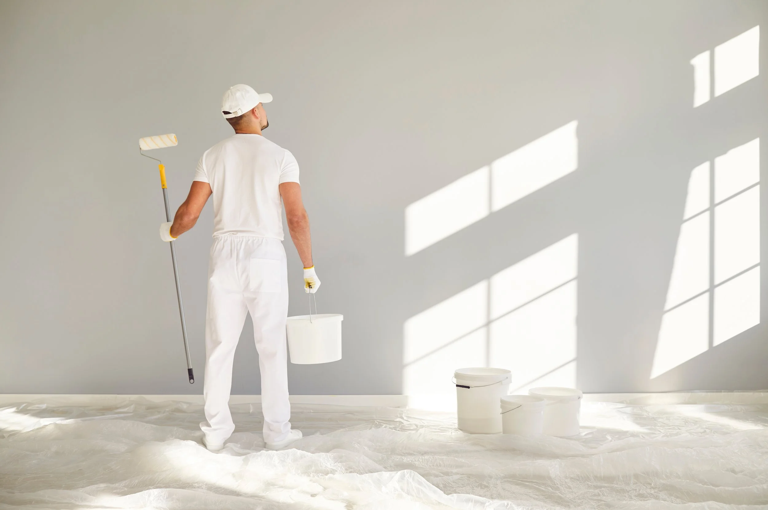 Person painting a wall white, holding a paint roller and a bucket, standing on a plastic sheet, with buckets on the floor and sunlight coming through window shadows.