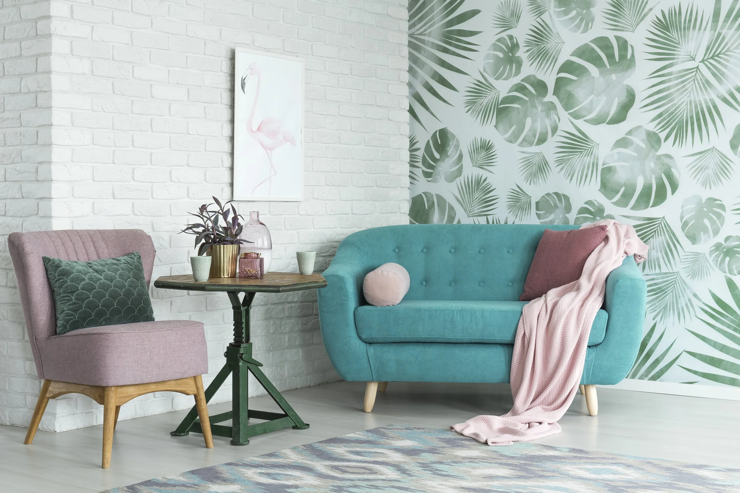 A living room corner with a pink armchair, a small wooden table, and a turquoise sofa. The wall has white brick and tropical leaf-patterned wallpaper. Decor includes a flamingo painting, plants, cups, and a pink blanket.