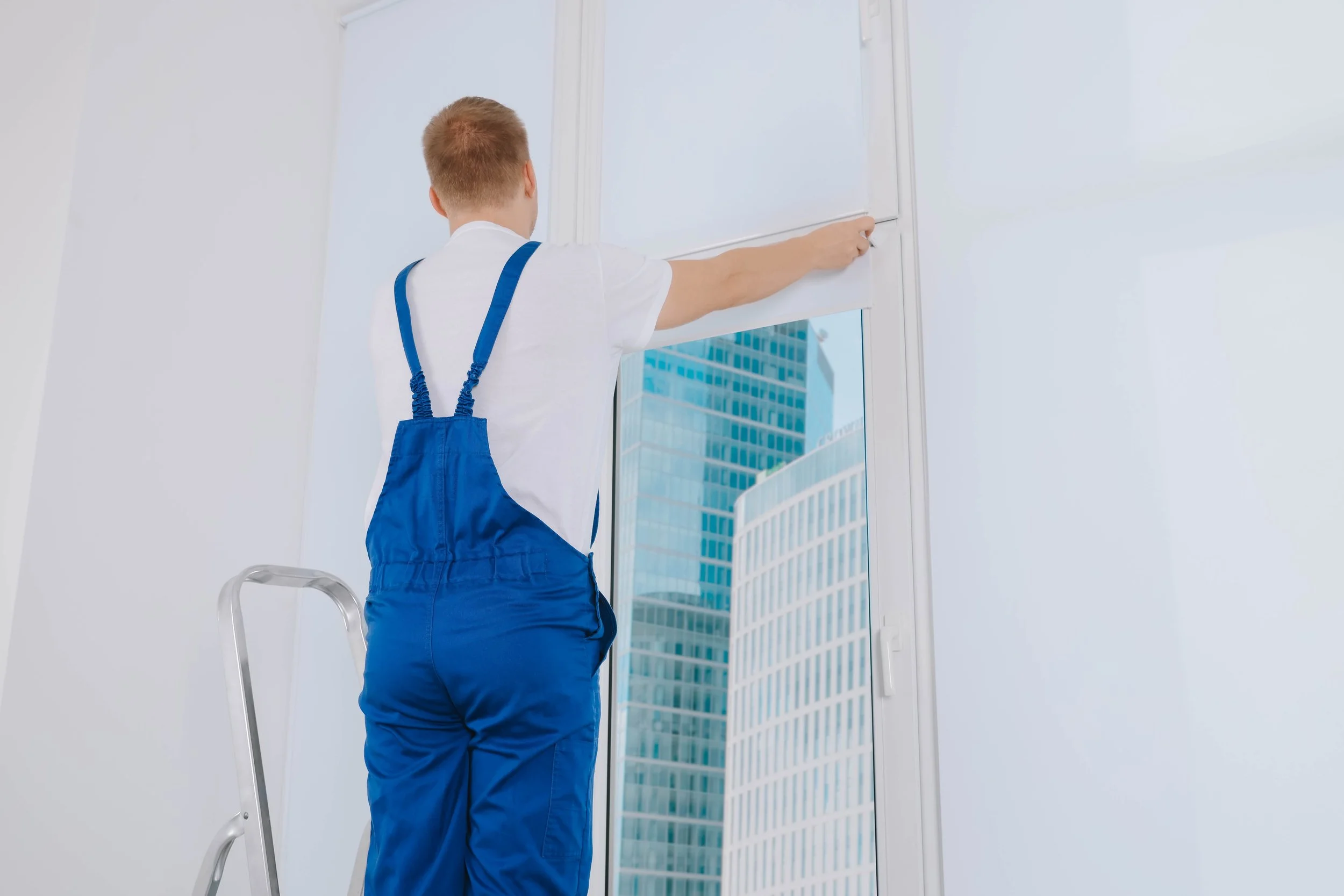 A man cleaning large window with a white roller. He is standing on a ladder, dressed in a white shirt and blue overalls, with modern city buildings visible through the window.