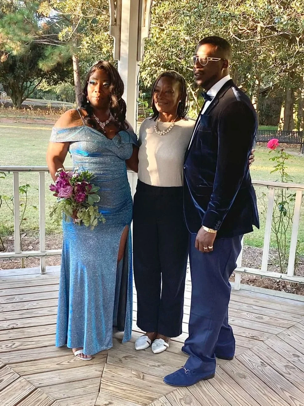 Three people standing on a wooden deck outdoors, dressed formally, two women and one man. The woman on the left is wearing a sparkly blue gown and holding a bouquet of flowers. The woman in the middle is wearing a white top and dark pants. The man on the right is dressed in a dark velvet tuxedo with sunglasses.