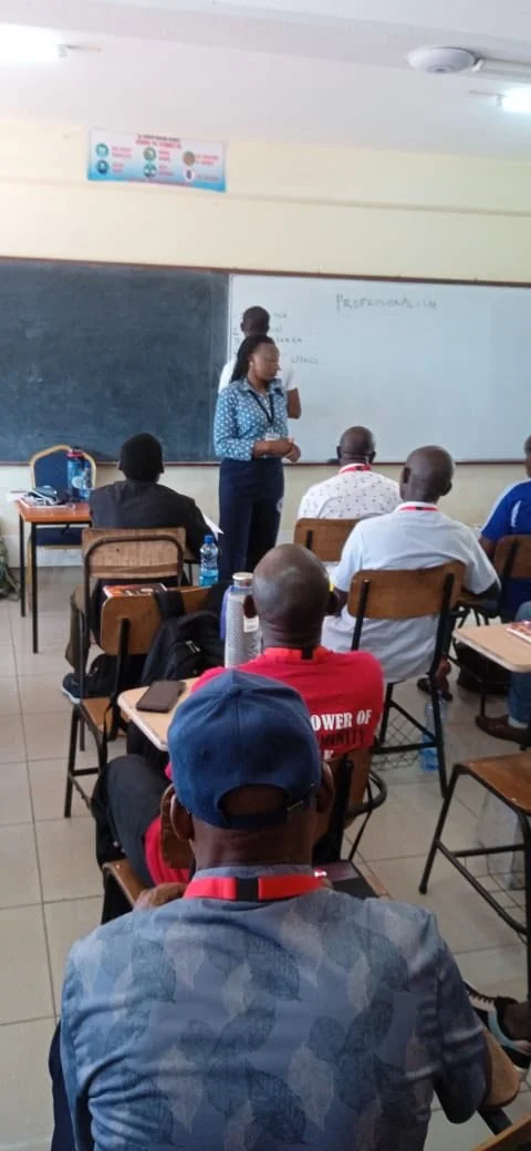A classroom with a female teacher standing at the blackboard, teaching students seated at desks. The students have notebooks and bottles on their desks, and the classroom has a white ceiling with a light fixture.