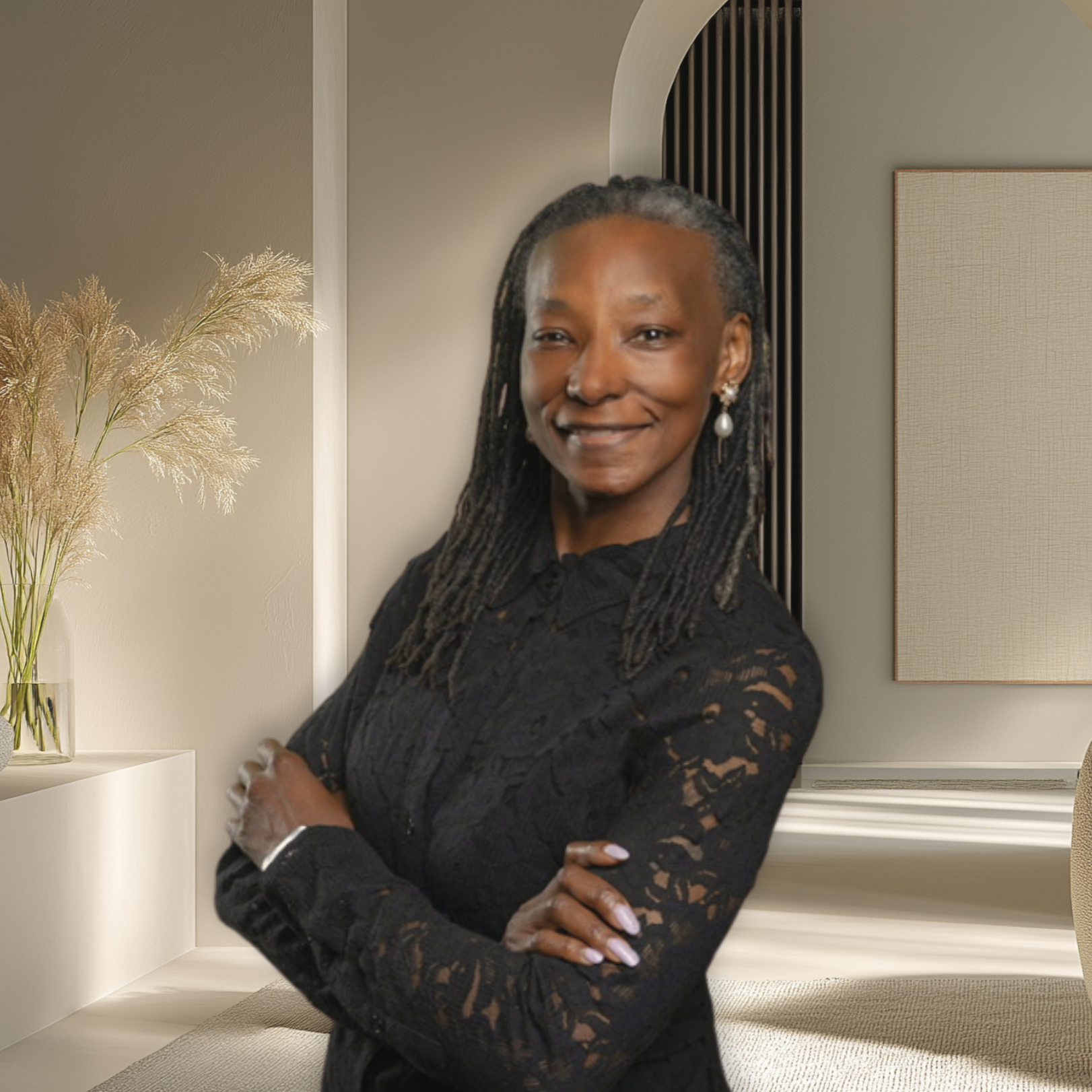 Portrait of a smiling woman with dark skin, long dreadlocks, in a black lace top, standing with folded arms in a modern, softly lit room with a vase of dried flowers and artwork on the wall.