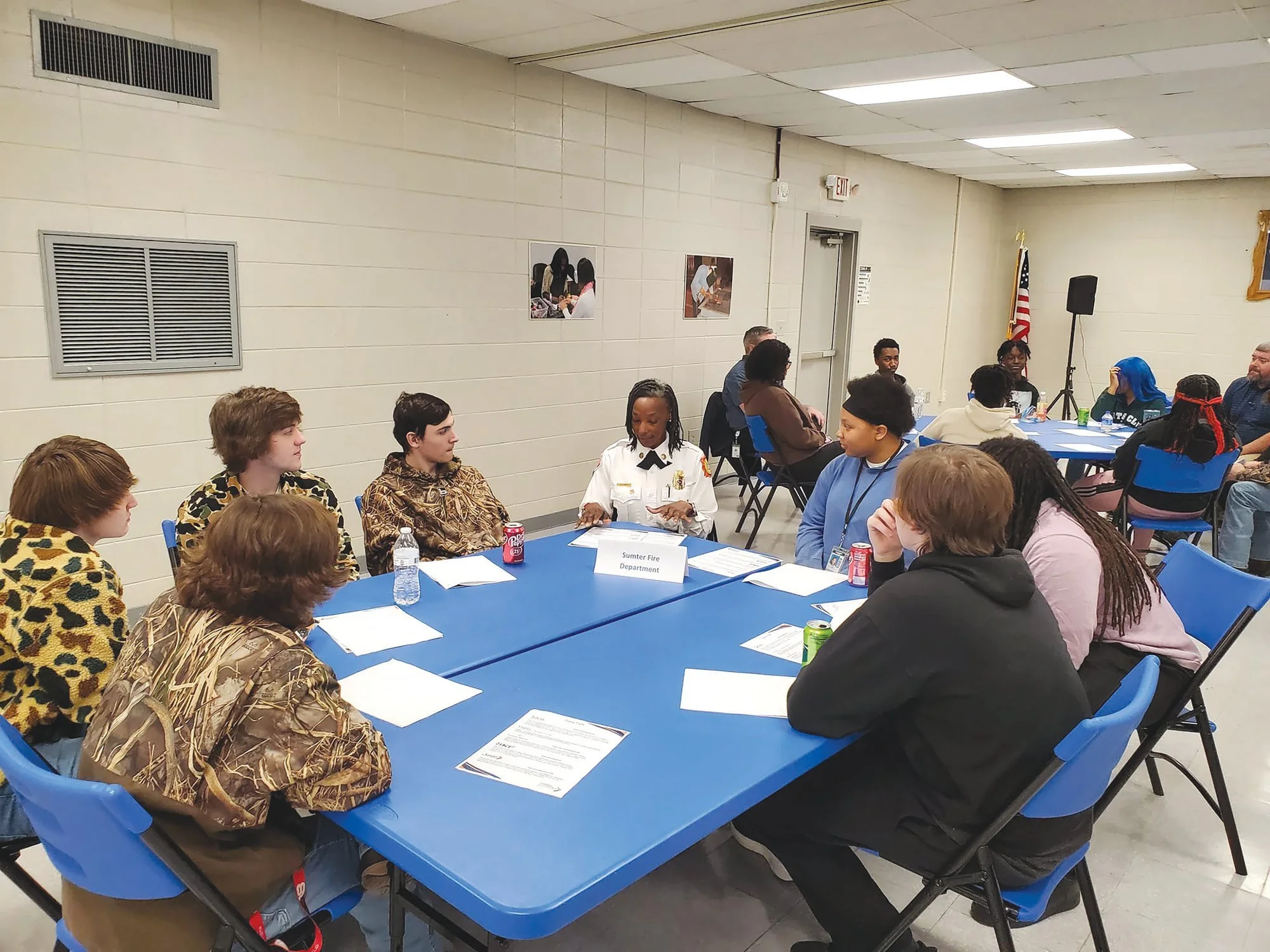 A group of people sitting around a blue table in a room, engaged in conversation. Some are wearing costumes, including animal print and camouflage. There are water bottles, cans of soda, and papers on the table. The room has beige walls, a white ceiling with fluorescent lights, and an American flag near a speaker. Two framed photos are hanging on the wall.