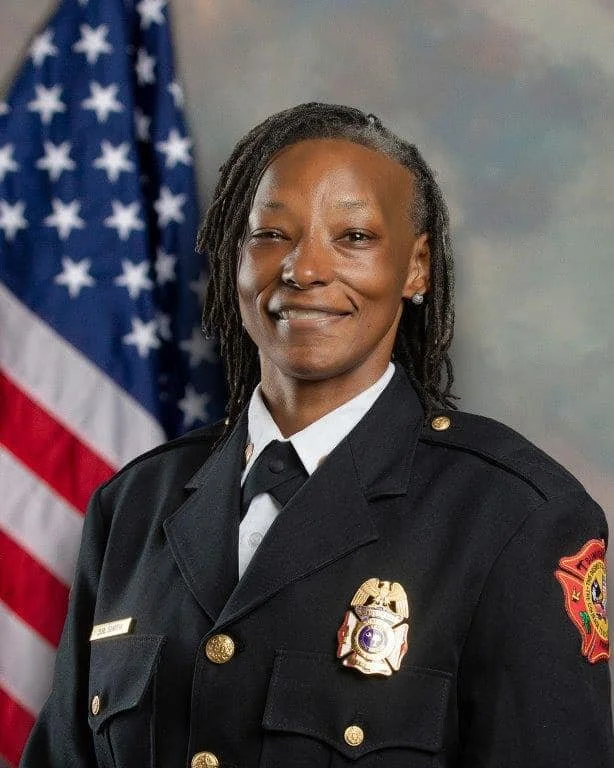 A female firefighter in uniform standing in front of an American flag, smiling.