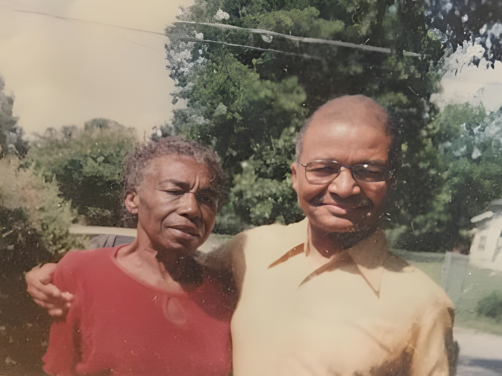 A woman and a man standing closely outdoors with trees in the background, smiling at the camera. The woman is wearing a red top and the man is wearing glasses and a yellow shirt.