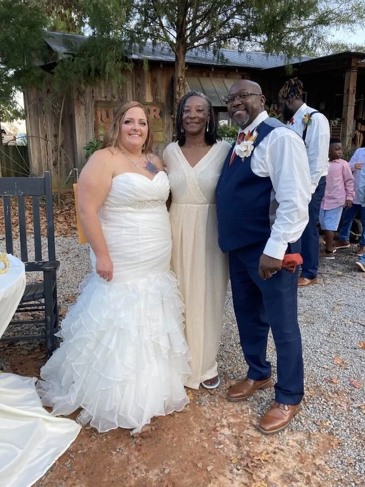Three people at a wedding, a bride in a white wedding gown, a woman in a cream dress, and a man in a blue vest and pants, smiling outdoors with a rustic wooden building and other guests in the background.