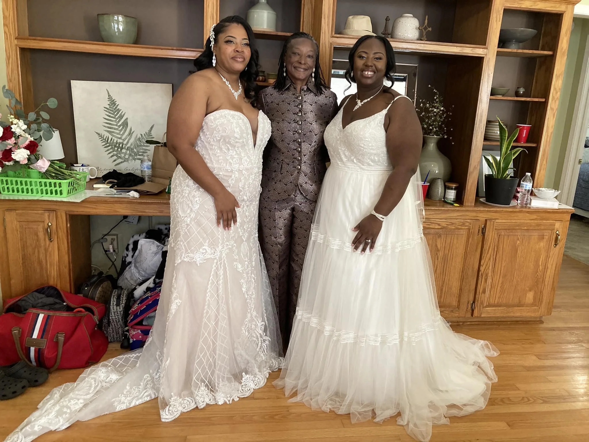 Three women standing inside a room. Two women are wearing white wedding dresses, and one woman is wearing a patterned jumpsuit. They are smiling and standing in front of a wooden shelf with decorative items. There are handbags, backpacks, and shoes on the floor near the shelf.