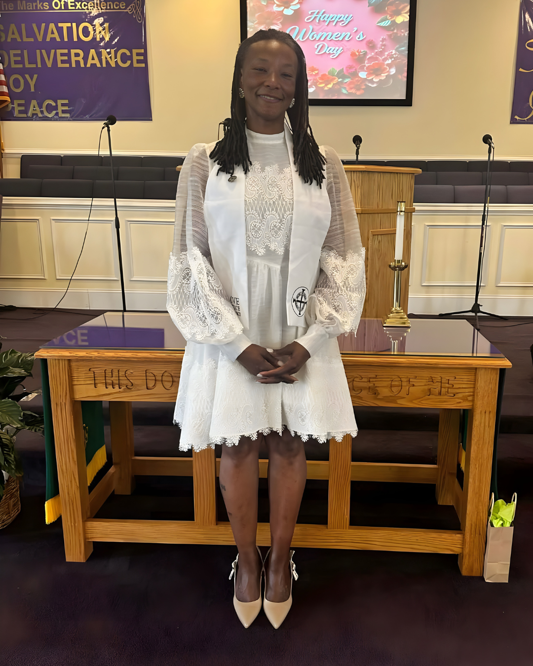 A woman in a white lace dress and beige high heels standing inside a church, smiling with her hands clasped in front of her. Behind her, there is a wooden lectern, a candle, and a large screen that reads 'Happy Women's Day'. The background features banners with religious text on the wall.