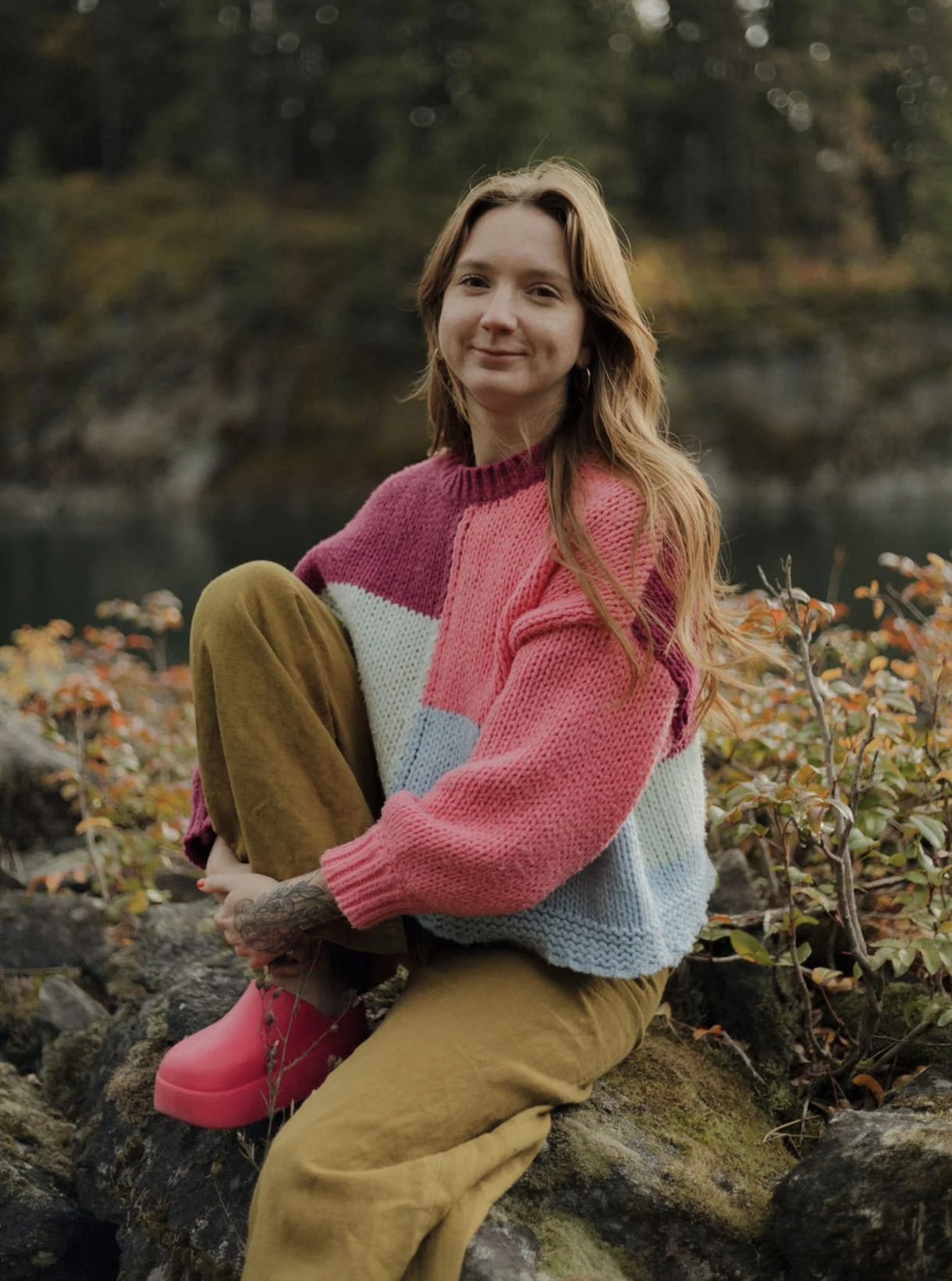 A woman with long, wavy reddish-brown hair sitting on rocks outdoors near a body of water, wearing a colorful knitted sweater and brown pants, with a tattoo on her left forearm, and pink boots.
