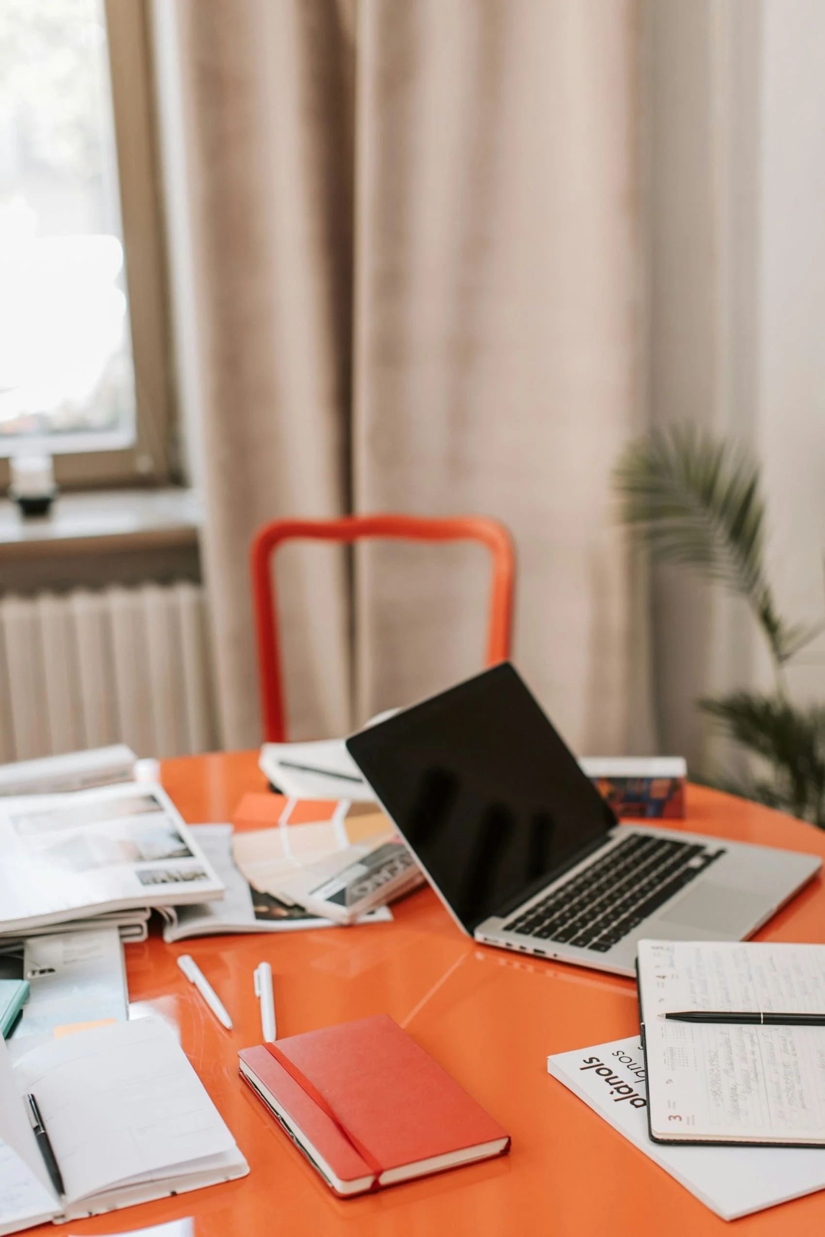 A cluttered orange table with notebooks, papers, and a laptop, in front of beige curtains and a window with natural light.
